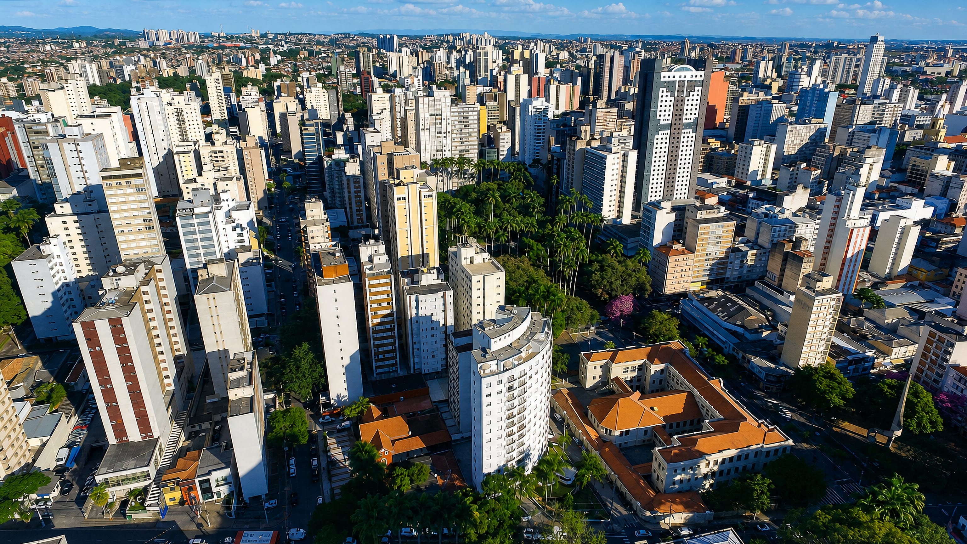 Aerial daytime view over Campinas, Brazil, showing dense apartment towers, palm-lined avenues and red-tiled rooftops.