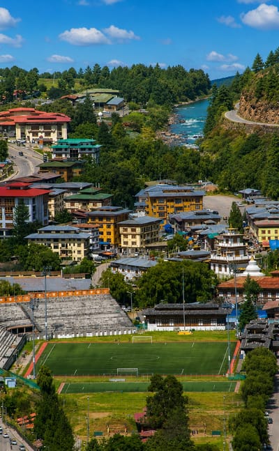 Thimphu in its forested valley with traditional Bhutanese buildings and a chorten beside a river and sports field