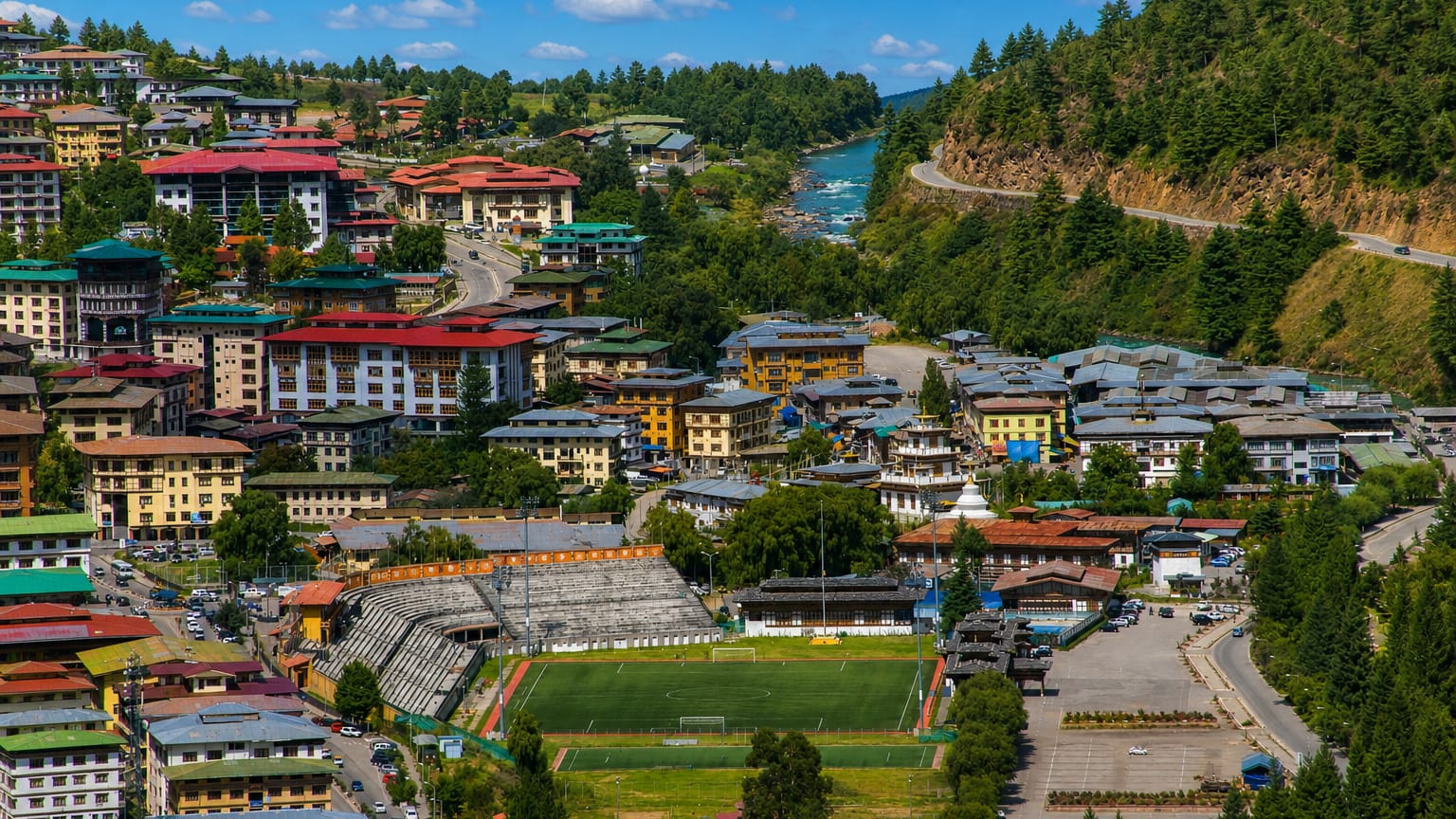 Thimphu in its forested valley with traditional Bhutanese buildings and a chorten beside a river and sports field