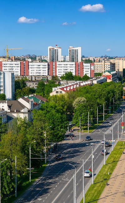 Brest, Belarus, a tree-lined avenue flanked by Soviet-era apartment blocks and parked cars under blue sky.