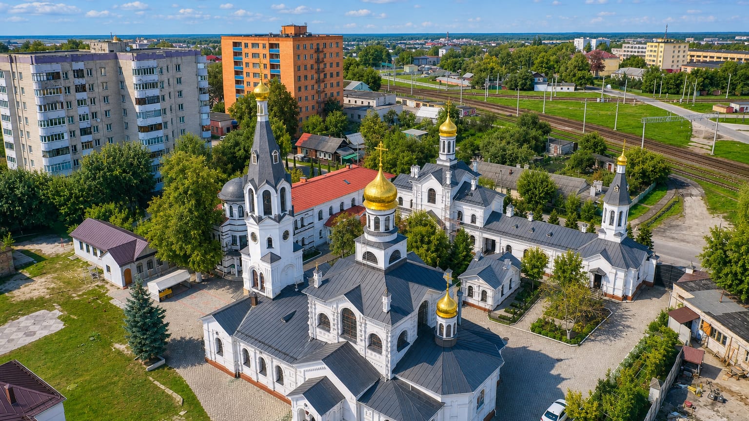 A white gold-domed Orthodox church among Soviet apartment blocks in Homyel', Belarus, beside railway tracks.