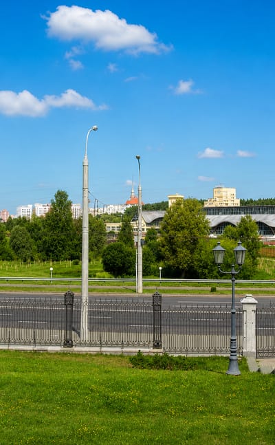 Wide boulevard with ornate lamp posts and an arena beyond greenery in the Pyershamayski district of Minsk