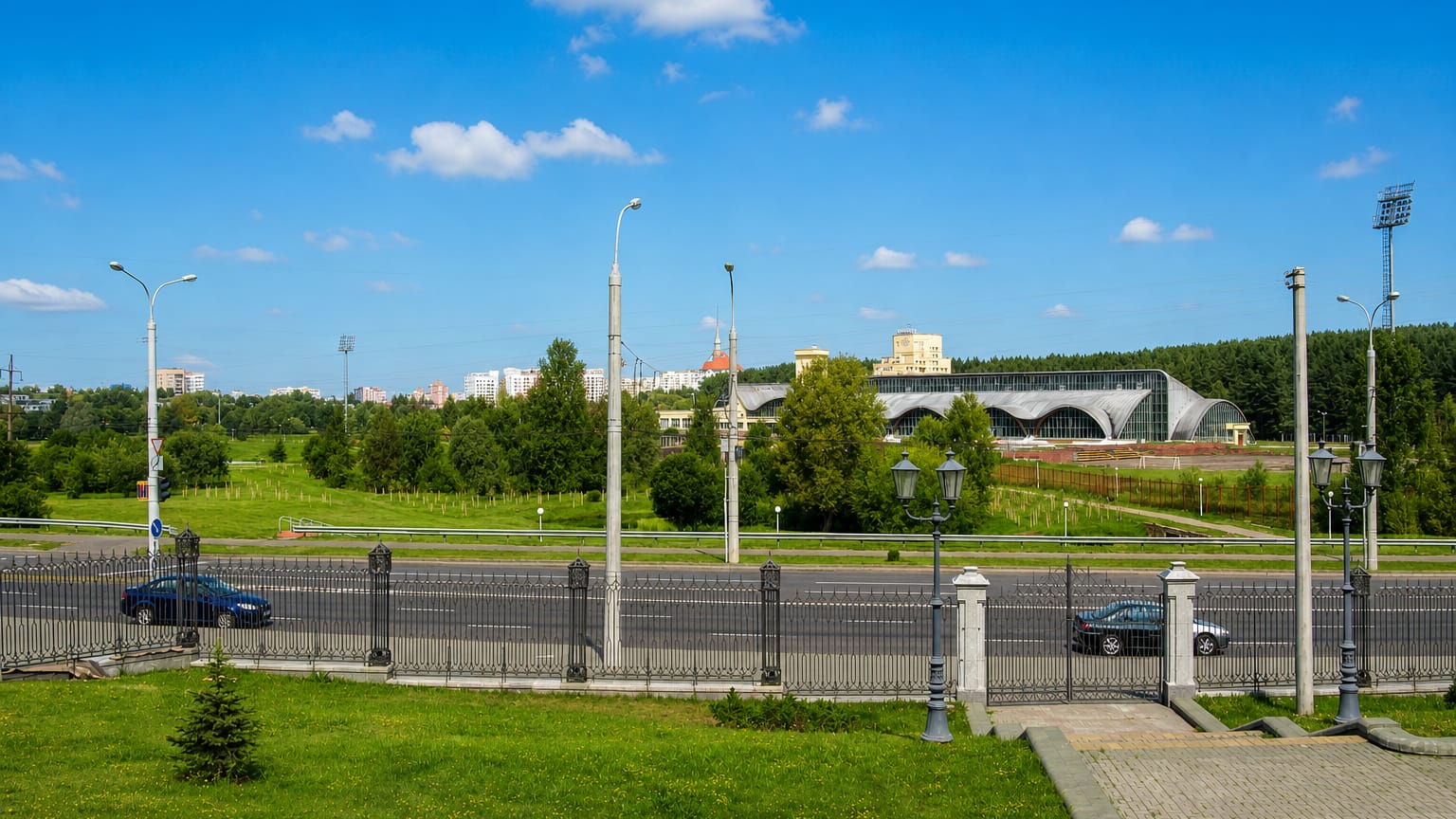 Wide boulevard with ornate lamp posts and an arena beyond greenery in the Pyershamayski district of Minsk
