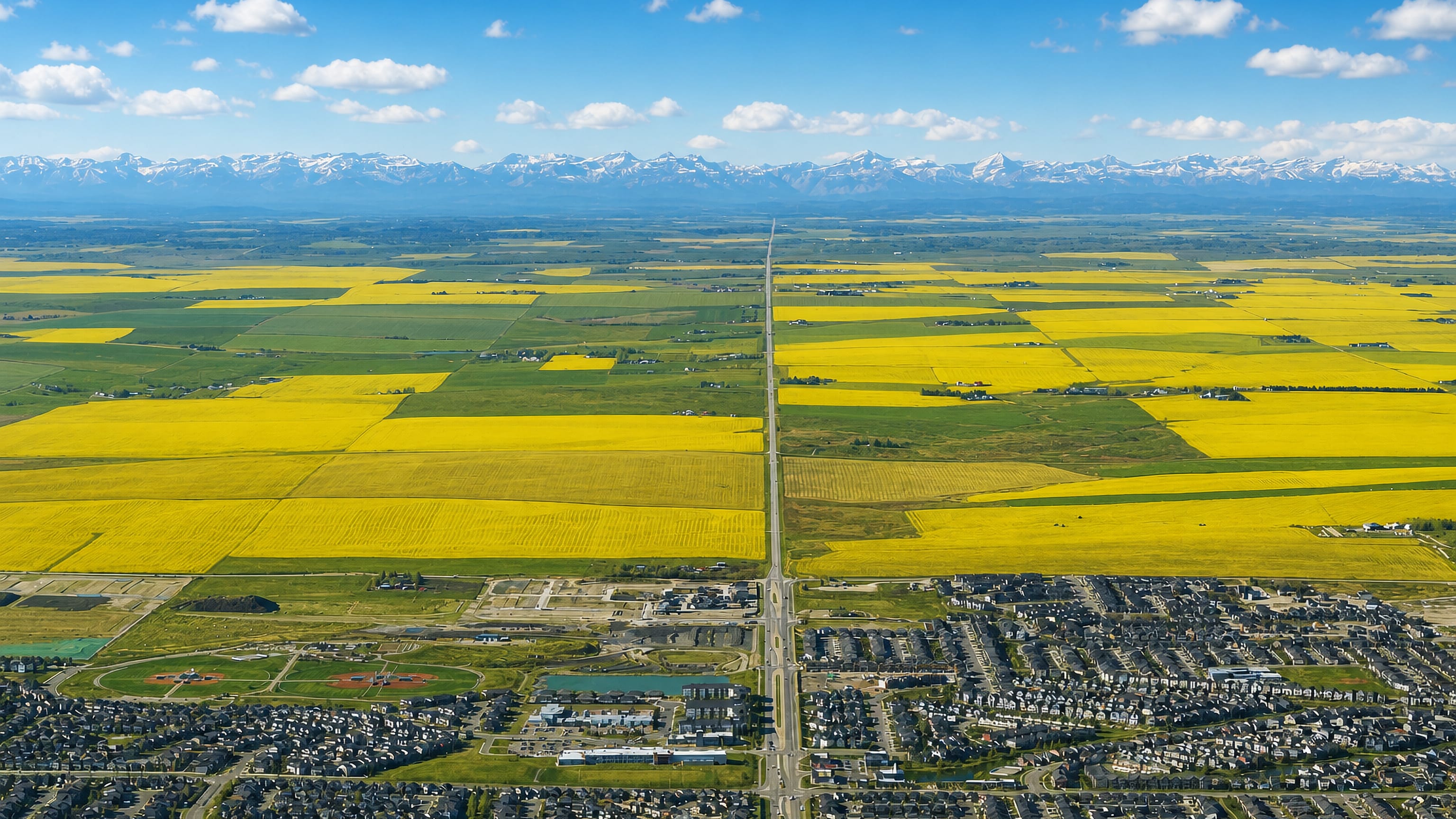 Airdrie, Alberta, spreading below yellow canola fields with the Rocky Mountains on the horizon.
