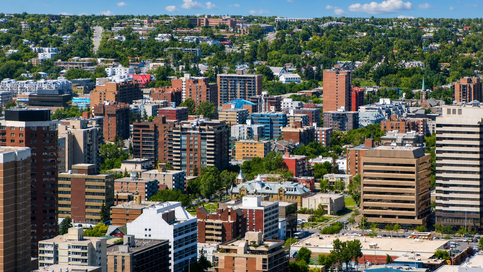 Dense inner-city Calgary district of mid-rise apartments and offices with tree-lined streets and hills on the horizon.