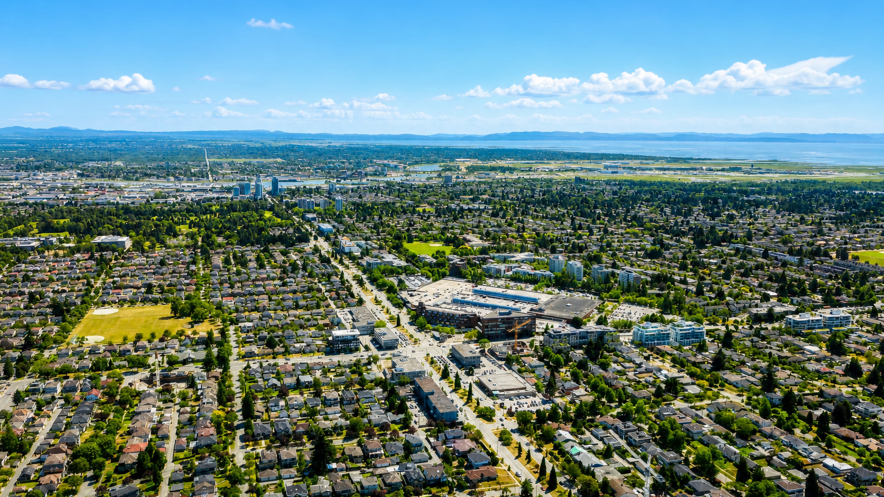 Aerial view of Oakridge in Vancouver, its shopping centre ringed by tree-lined streets of houses, sea beyond