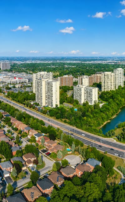 Aerial view of Brampton's apartment tower clusters and tree-lined suburban homes beside a highway and pond.