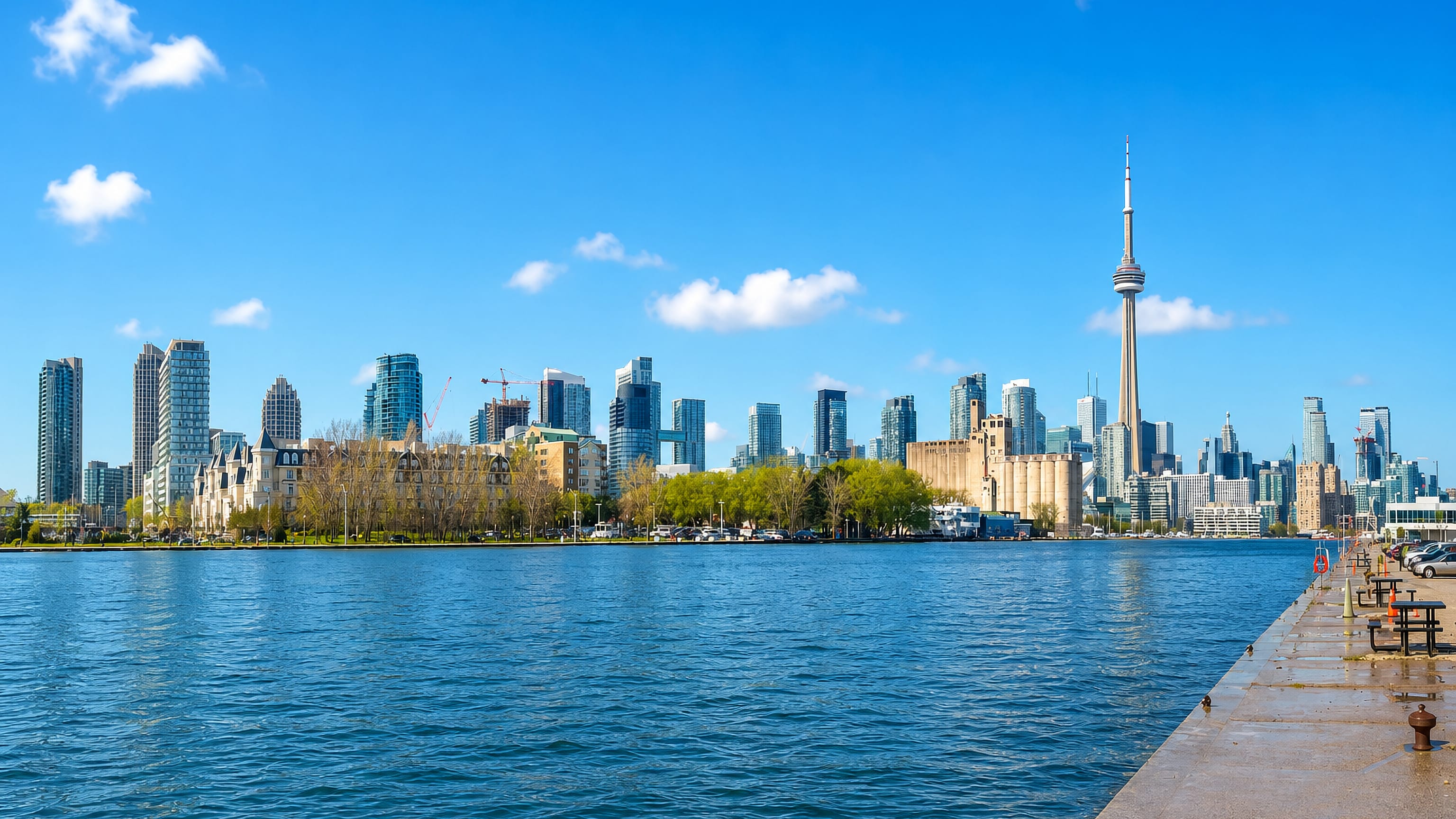 Toronto's downtown skyline with the CN Tower seen across Lake Ontario from a waterfront pier under a blue sky.