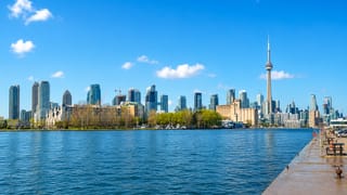 Toronto's downtown skyline with the CN Tower seen across Lake Ontario from a waterfront pier under a blue sky.