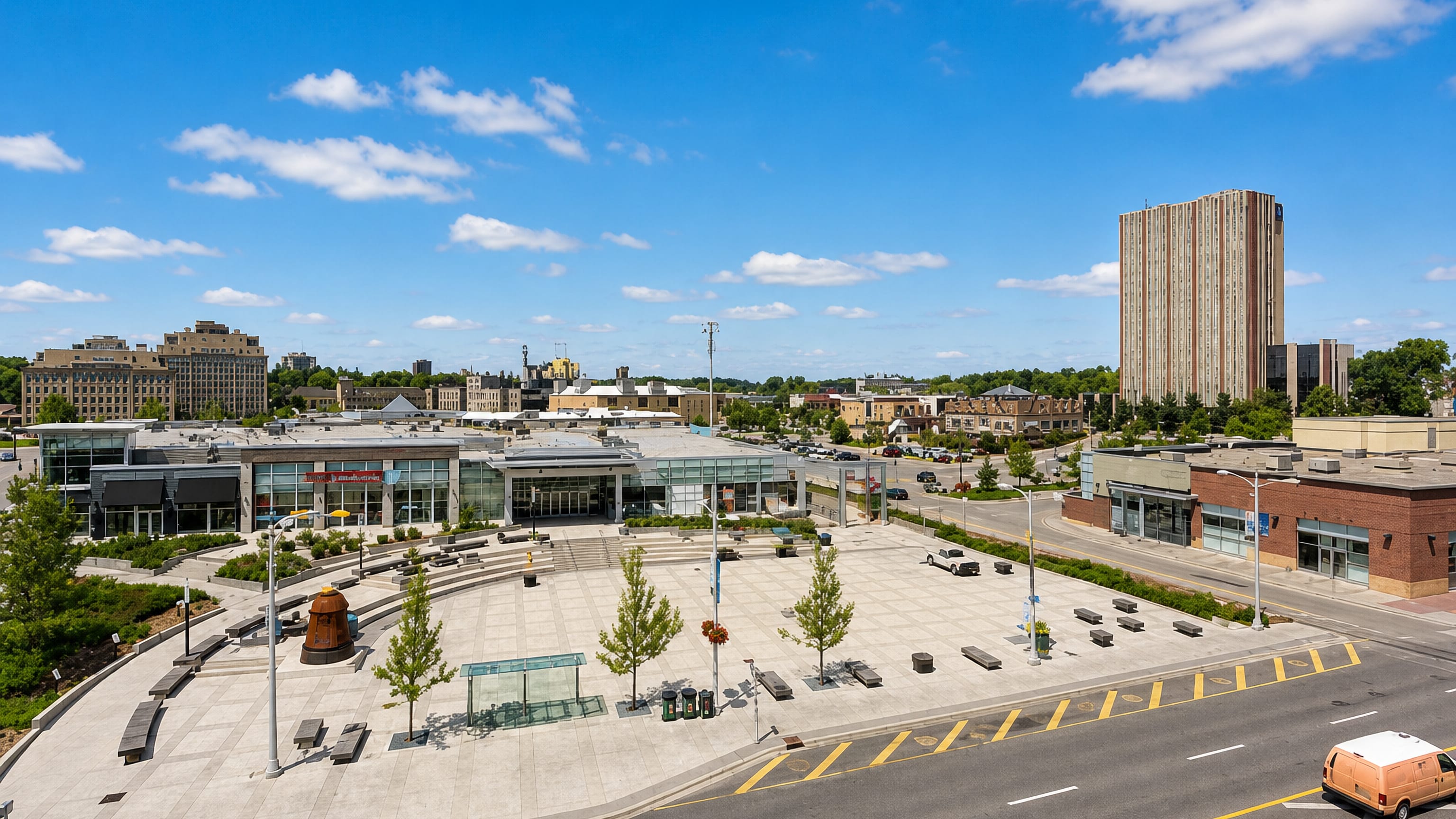 Uptown Waterloo public square with paved plaza, low glass storefronts and a tower block under blue sky, Ontario.