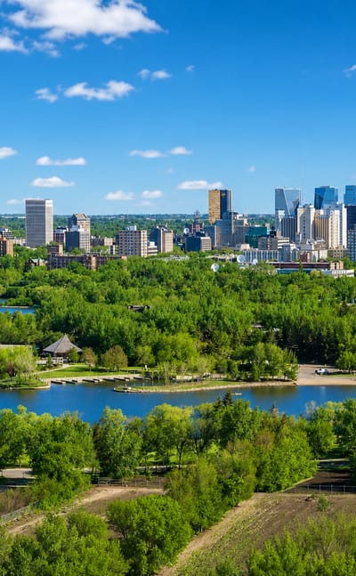 Downtown Regina rises beyond the green shoreline of Wascana Lake under a blue prairie sky.