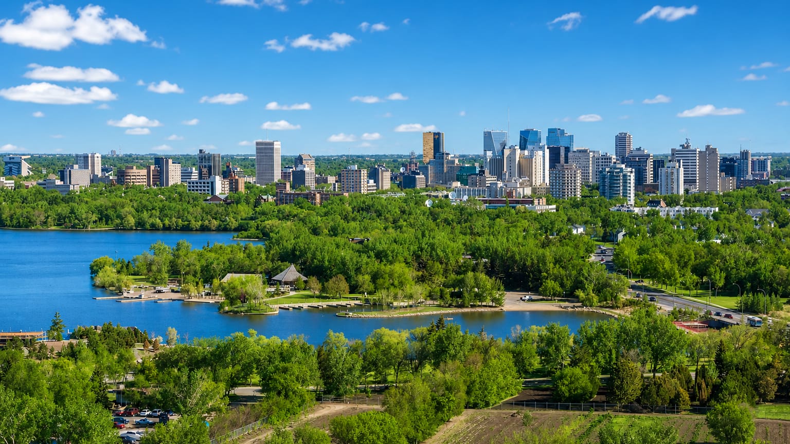Downtown Regina rises beyond the green shoreline of Wascana Lake under a blue prairie sky.