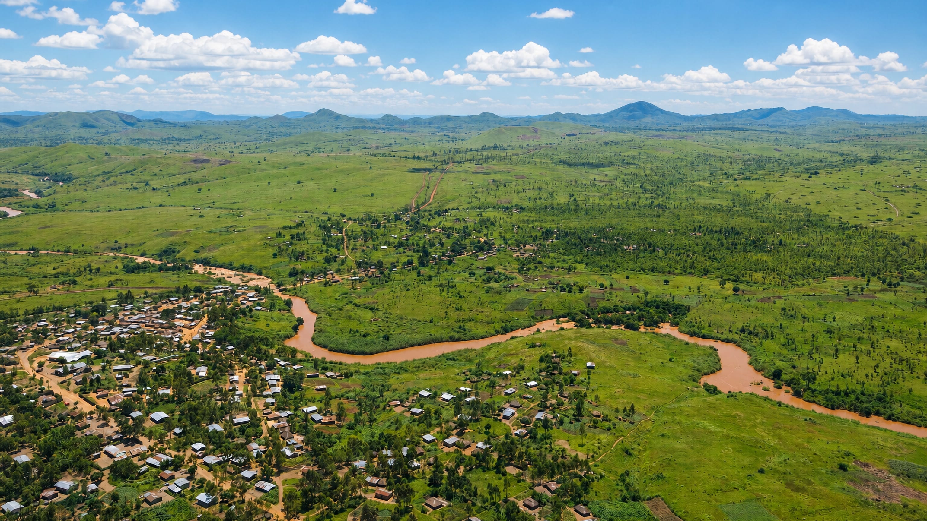 Aerial view over Bunia, DR Congo, with tin-roofed homes, red-earth roads and a muddy river winding through green hills.