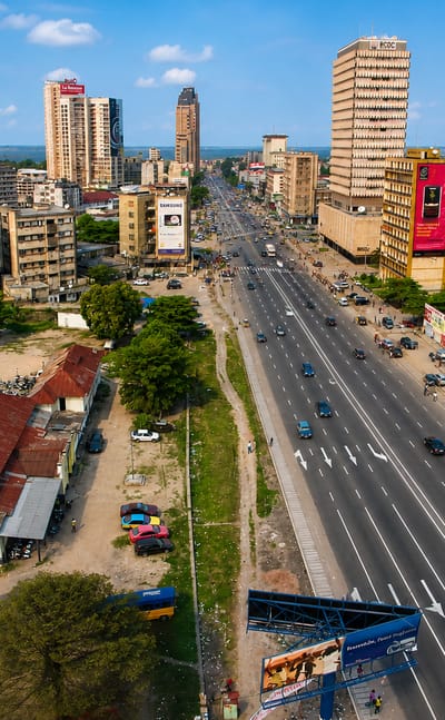 Kinshasa's Boulevard du 30 Juin with mid-rise towers, billboards and traffic under a partly cloudy sky.