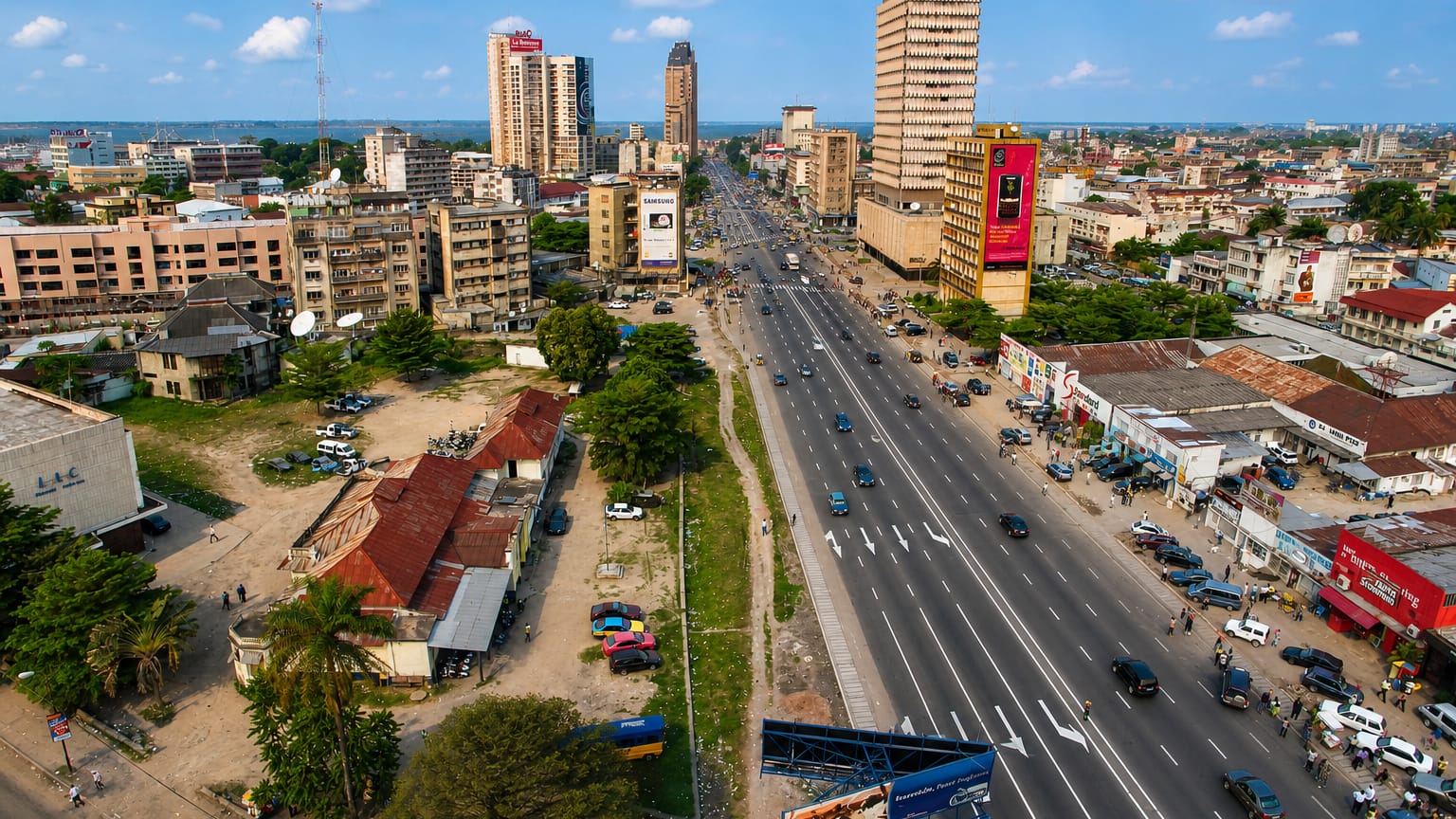 Kinshasa's Boulevard du 30 Juin with mid-rise towers, billboards and traffic under a partly cloudy sky.