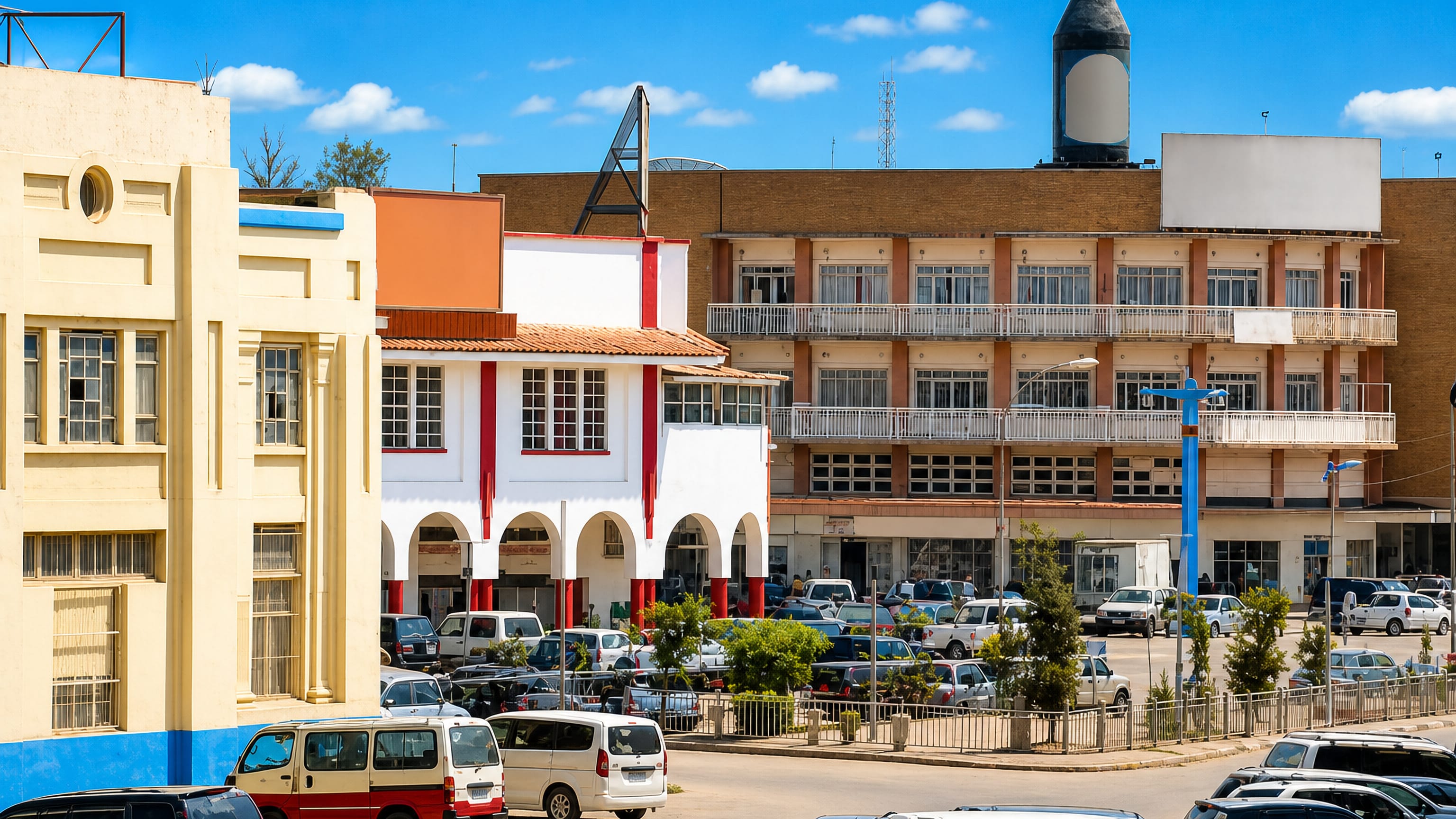 Sunlit downtown Lubumbashi street with mid-century arcaded shopfronts, balconied apartment blocks and parked cars.
