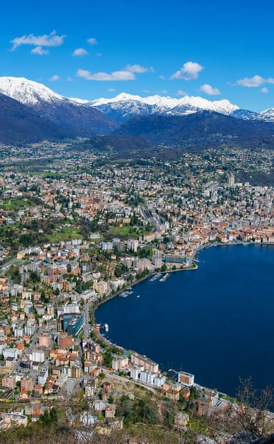 Elevated view of Lugano, Switzerland, wrapping around its lake beneath snow-capped Alpine peaks.