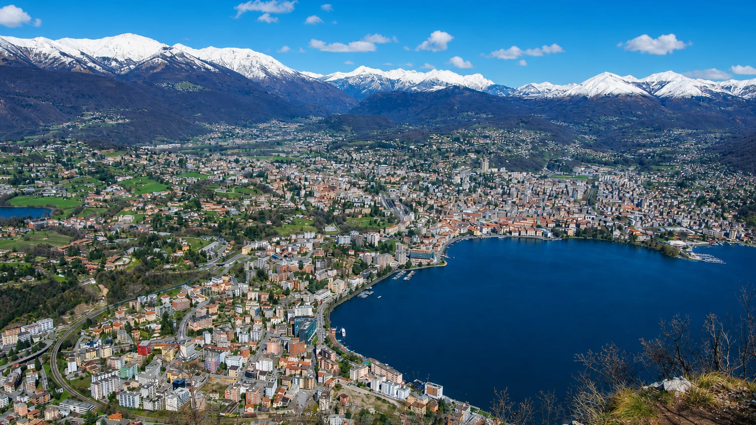 Elevated view of Lugano, Switzerland, wrapping around its lake beneath snow-capped Alpine peaks.