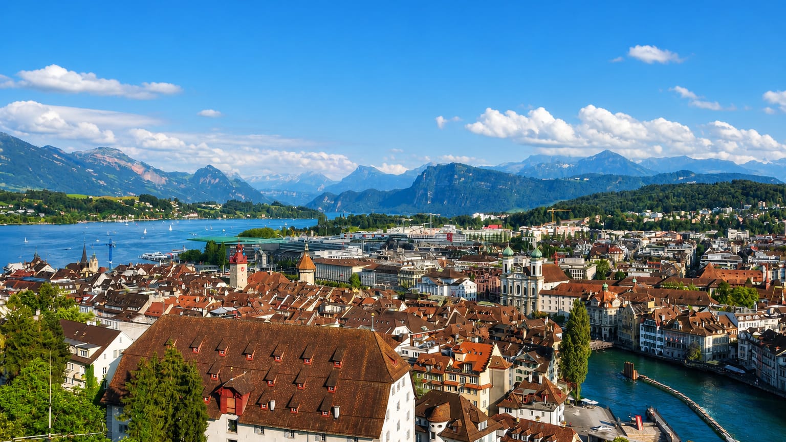 Panorama of Lucerne, Switzerland, over the old town's red rooftops toward the lake and snow-tinged Alps.