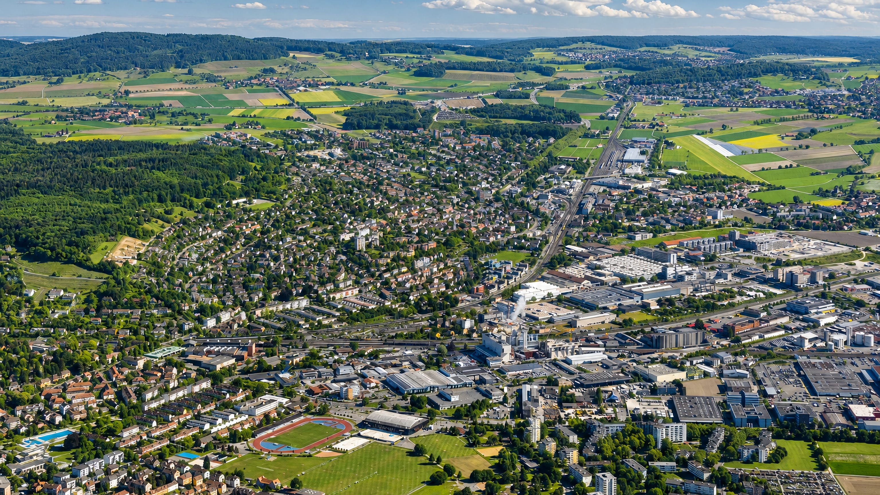 Aerial view over Oberwinterthur in Winterthur, its housing, sports track and industry ringed by farmland and hills.