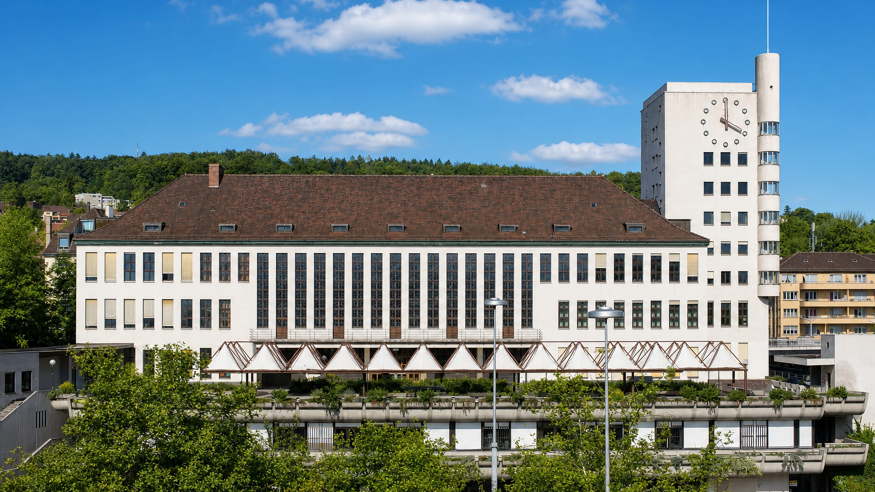 A white modernist building with a clock tower in Wipkingen, Zurich Kreis 10, below the wooded Kaeferberg hillside.