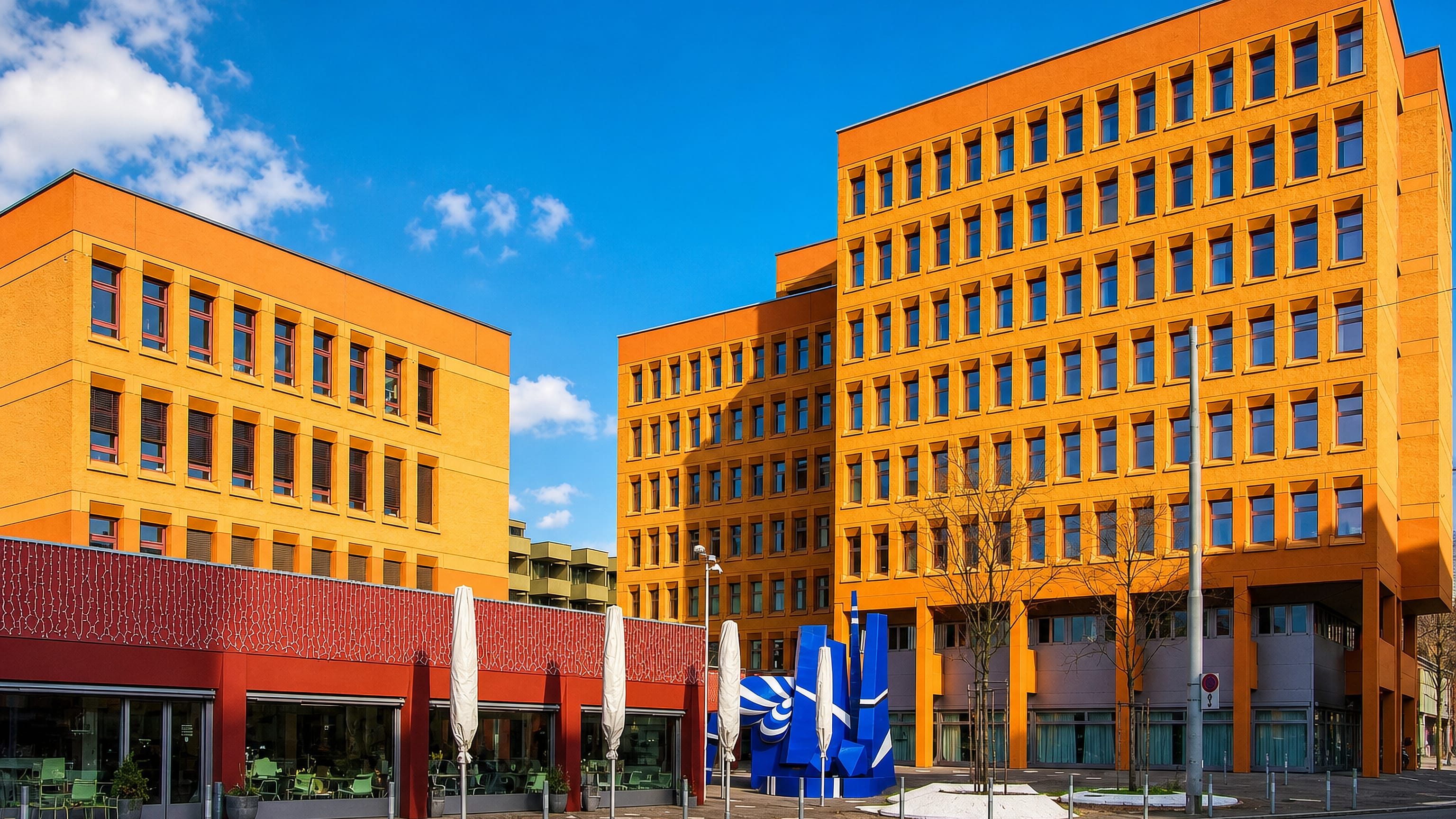 Orange-clad modern office blocks and a blue steel sculpture along a street in Zurich's Oerlikon quarter.