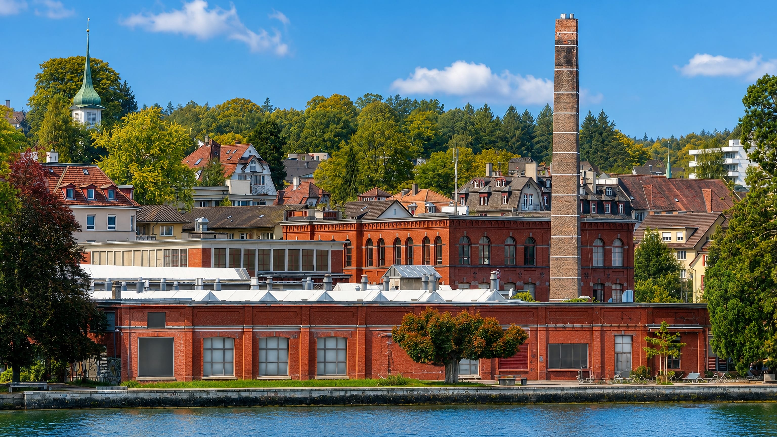 Red-brick lakeside factory with a tall chimney below villas and woods at Wollishofen on Lake Zurich
