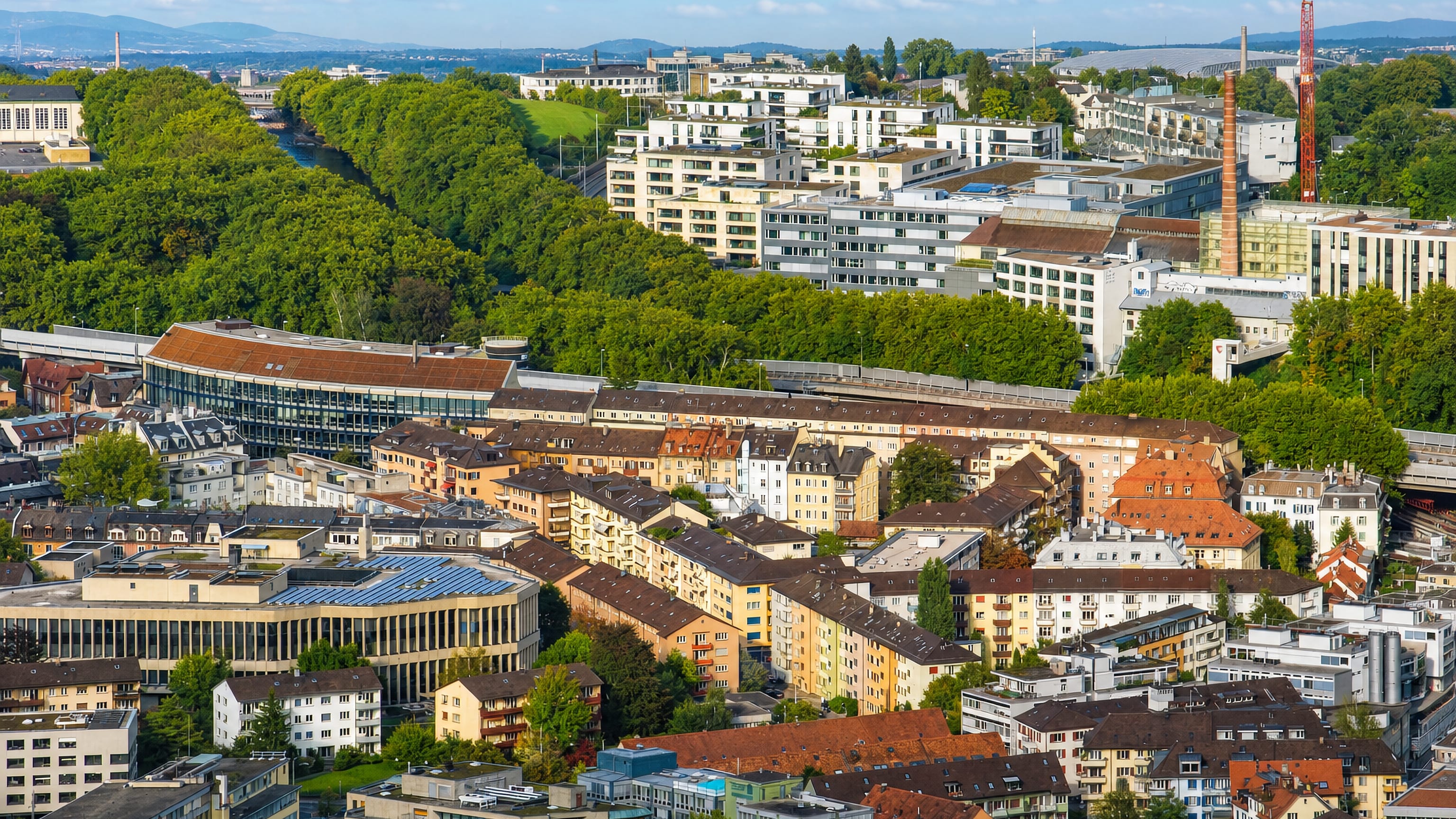 Aerial view of Zürich Kreis 3, with tiled rooftops, office blocks and the wooded Sihl river corridor beyond.
