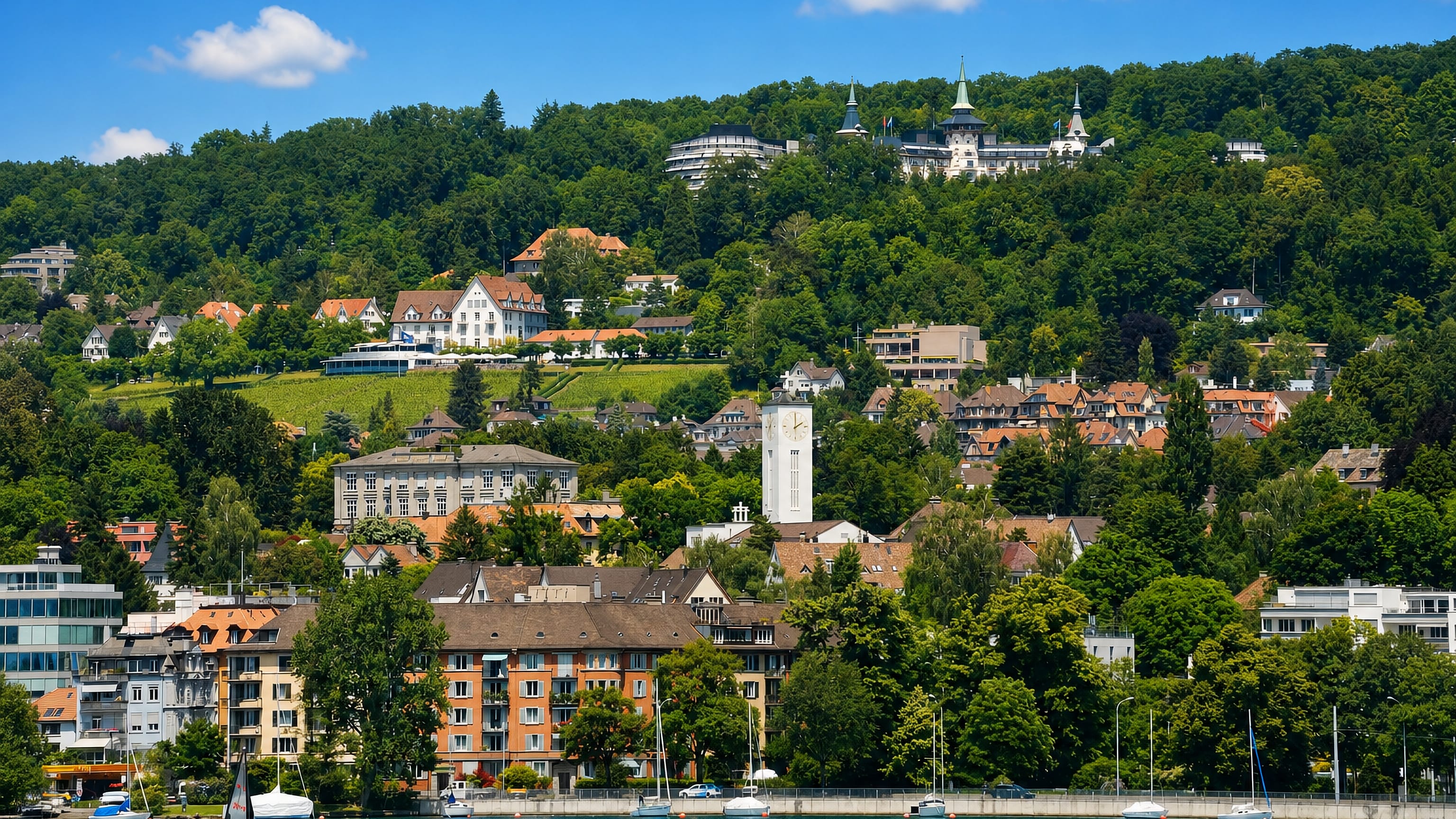 Lake Zurich shoreline below wooded slopes and a turreted hilltop hotel in Zurich's Kreis 8.