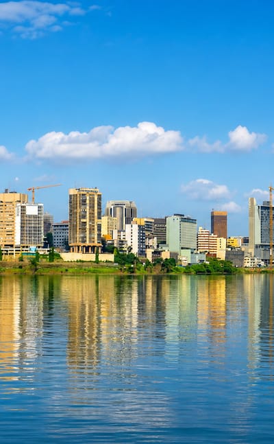 Abidjan's Plateau high-rise skyline in daylight, mirrored in the Ébrié Lagoon under a blue sky.
