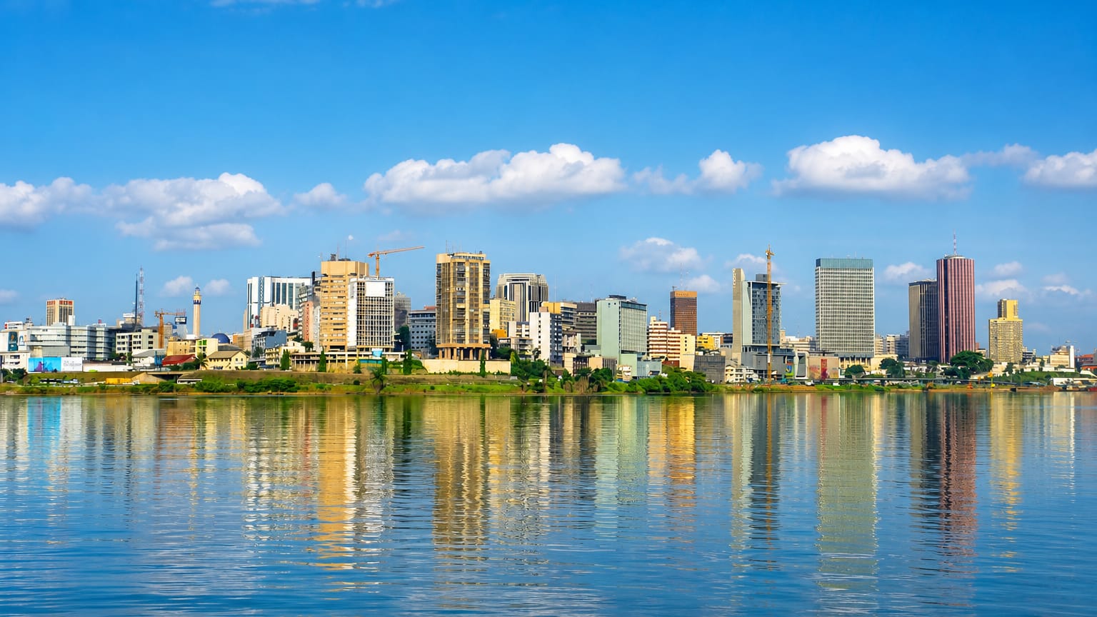 Abidjan's Plateau high-rise skyline in daylight, mirrored in the Ébrié Lagoon under a blue sky.