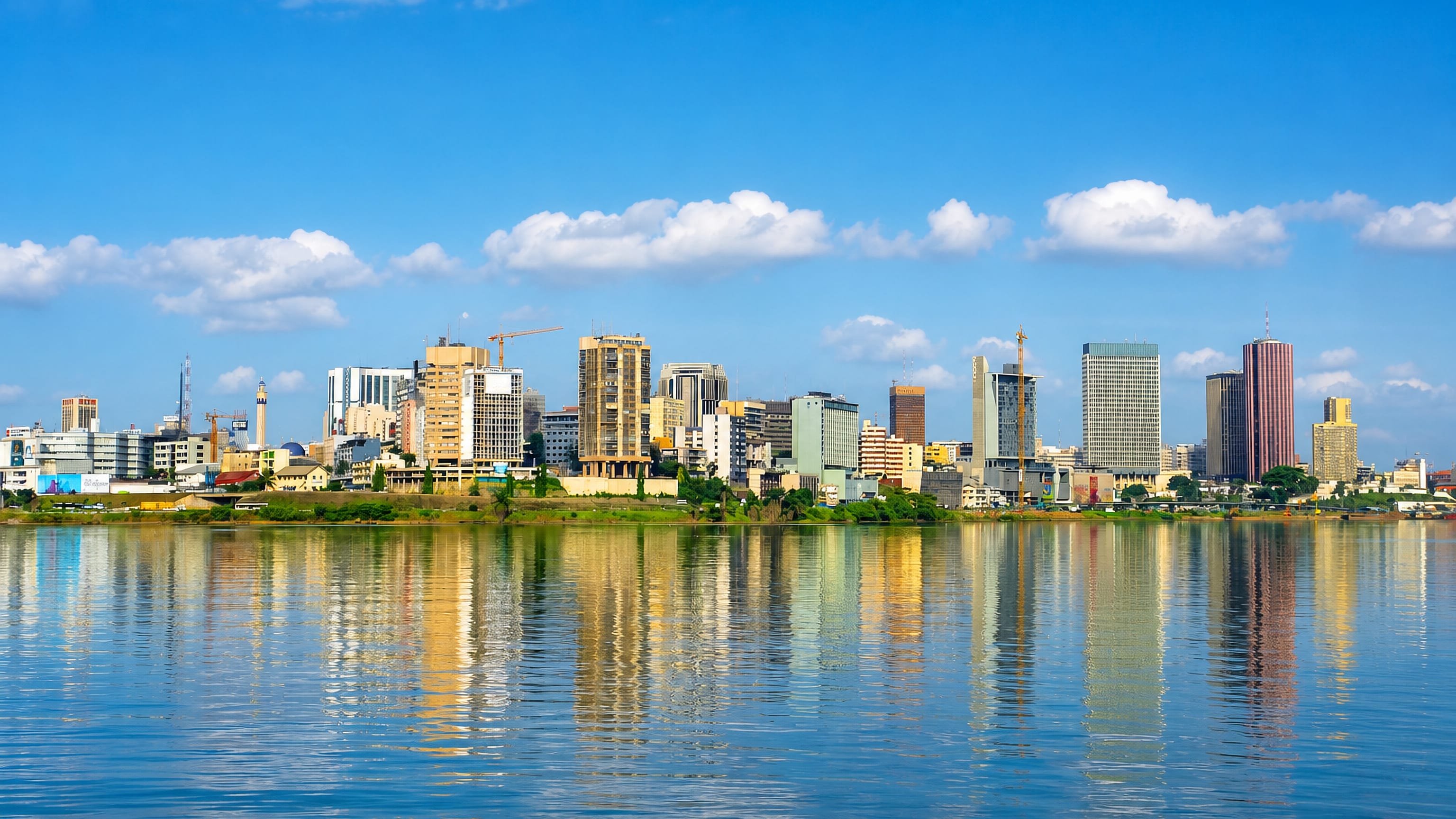Abidjan's Plateau high-rise skyline in daylight, mirrored in the Ébrié Lagoon under a blue sky.