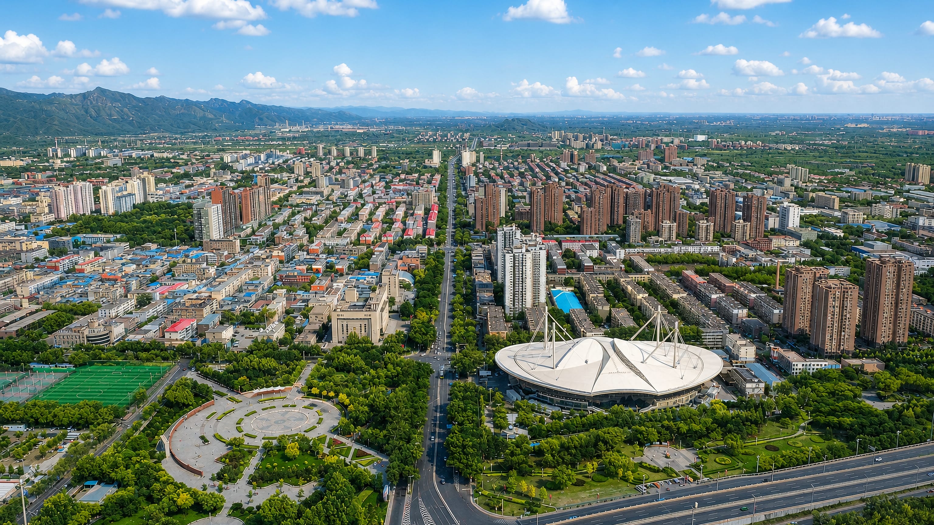 Aerial over Changping District, Beijing, with a tensile-roofed arena, a wide boulevard and mountains beyond