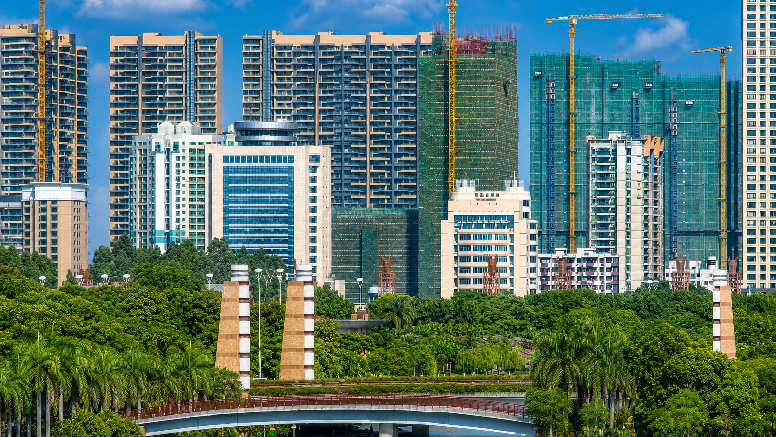 High-rise towers and construction cranes above a palm-lined park and canal bridge in Nanhai, Foshan