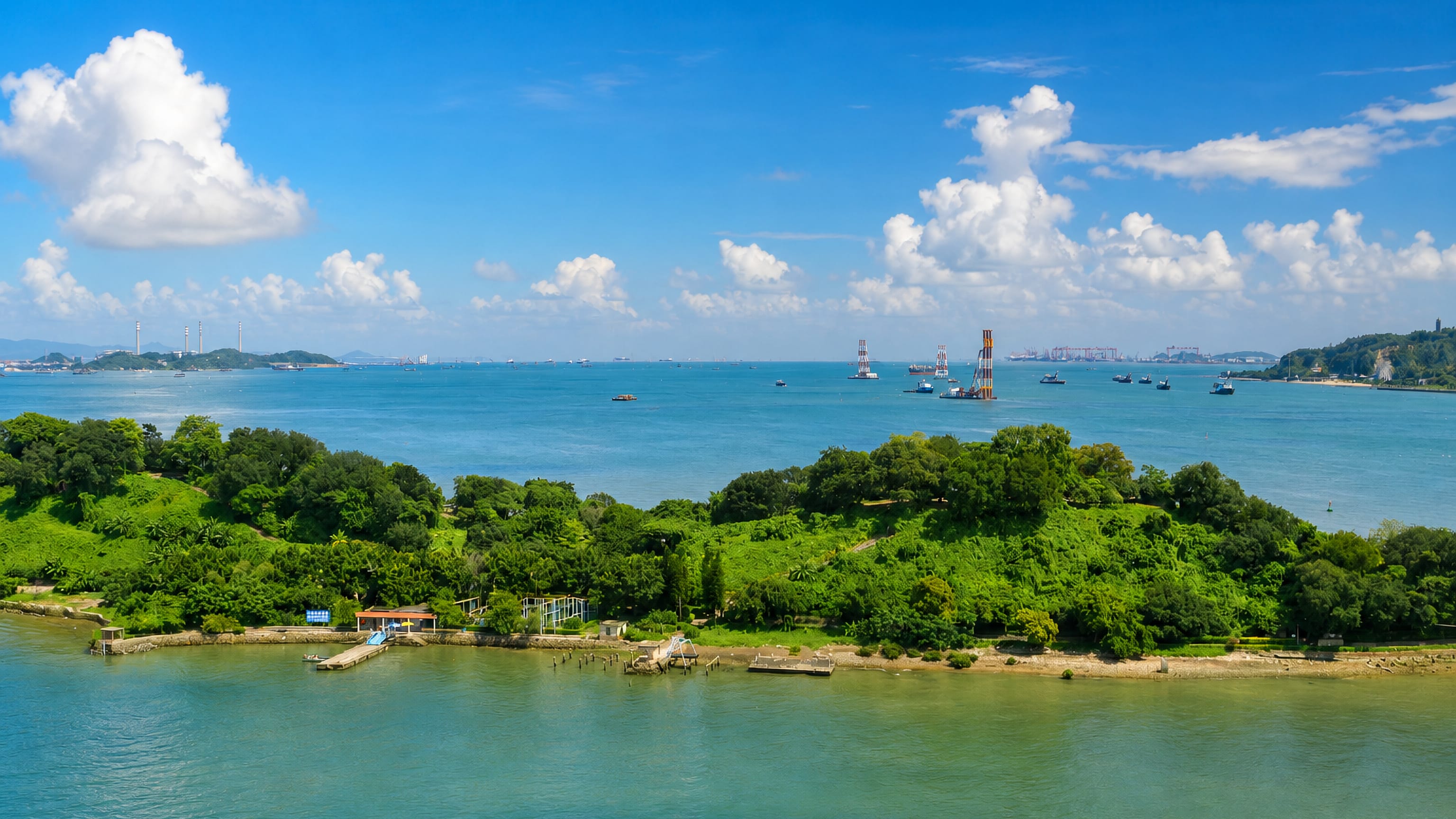 Wooded islet in the Pearl River estuary off Nansha District, Guangzhou, with jack-up rigs and port cranes beyond