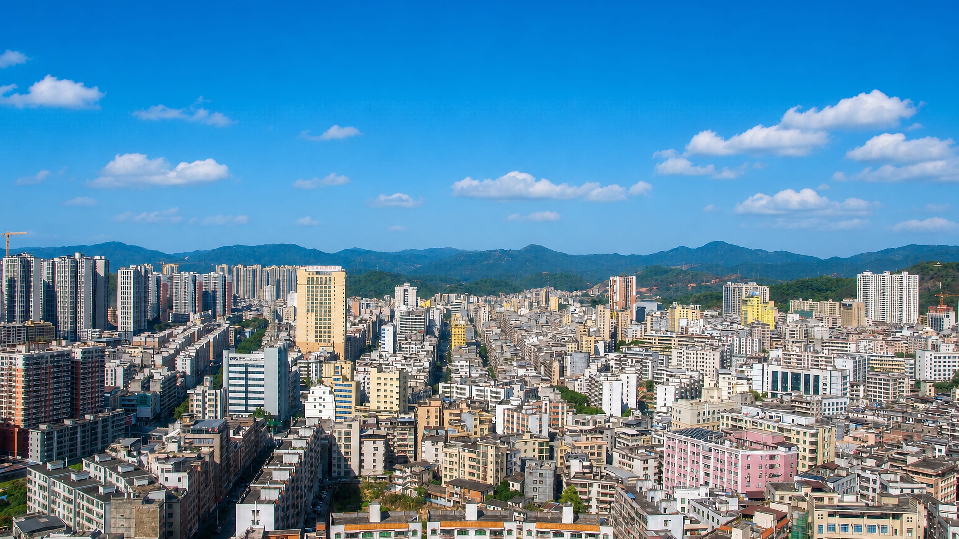 Aerial daytime view over Maoming's dense high-rise and low-rise districts in Guangdong, with green hills beyond.