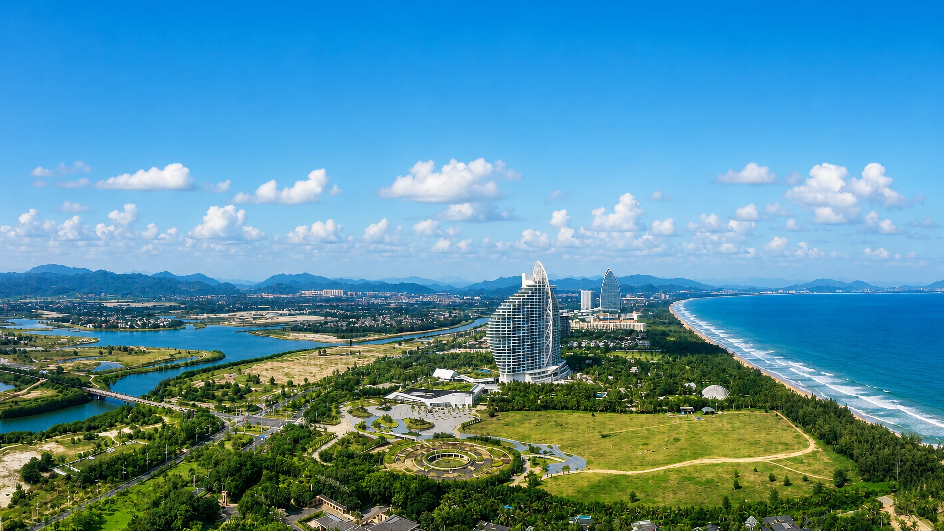 Aerial view of Sanya's Hainan coastline, with a sail-shaped resort tower, river mouth, palms and a long sandy beach.