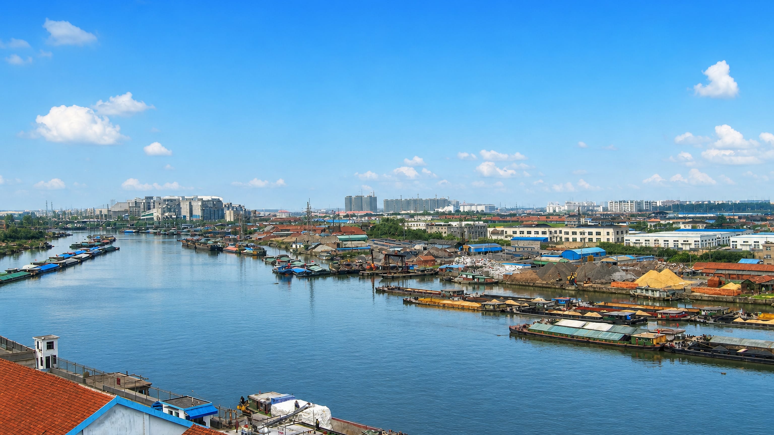Cargo barges and sand yards line a wide canal in Yancheng, Jiangsu, with the city's low skyline behind.