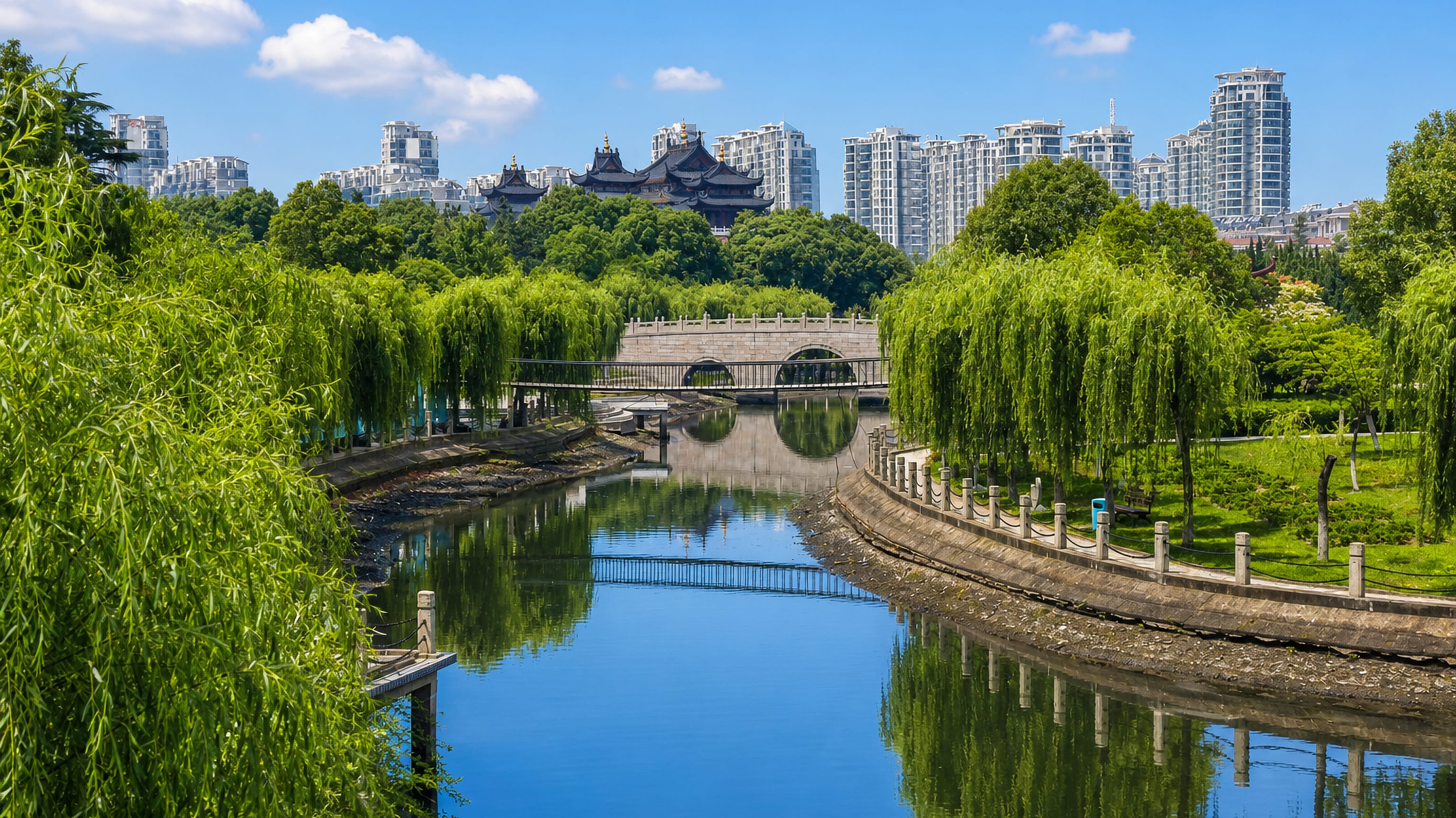 Willow-lined canal and stone arch bridge in Zhangjiagang, with temple roofs and high-rises beyond.