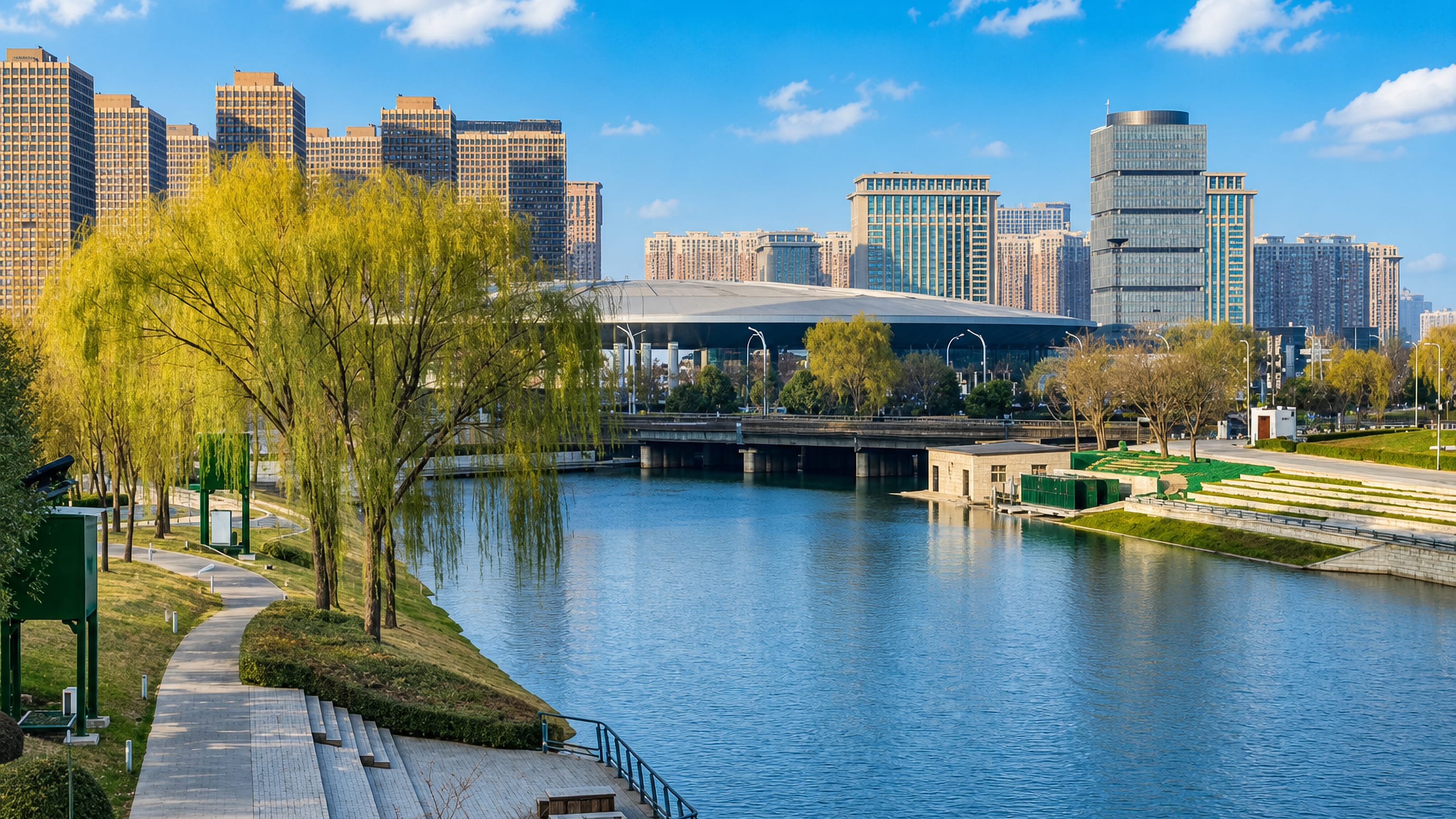 Willow-lined Fen River waterfront park in Taiyuan backed by a domed arena and high-rise towers.