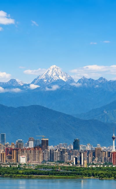 Chengdu's high-rise skyline and TV tower with snow-capped mountains rising behind under a blue sky.
