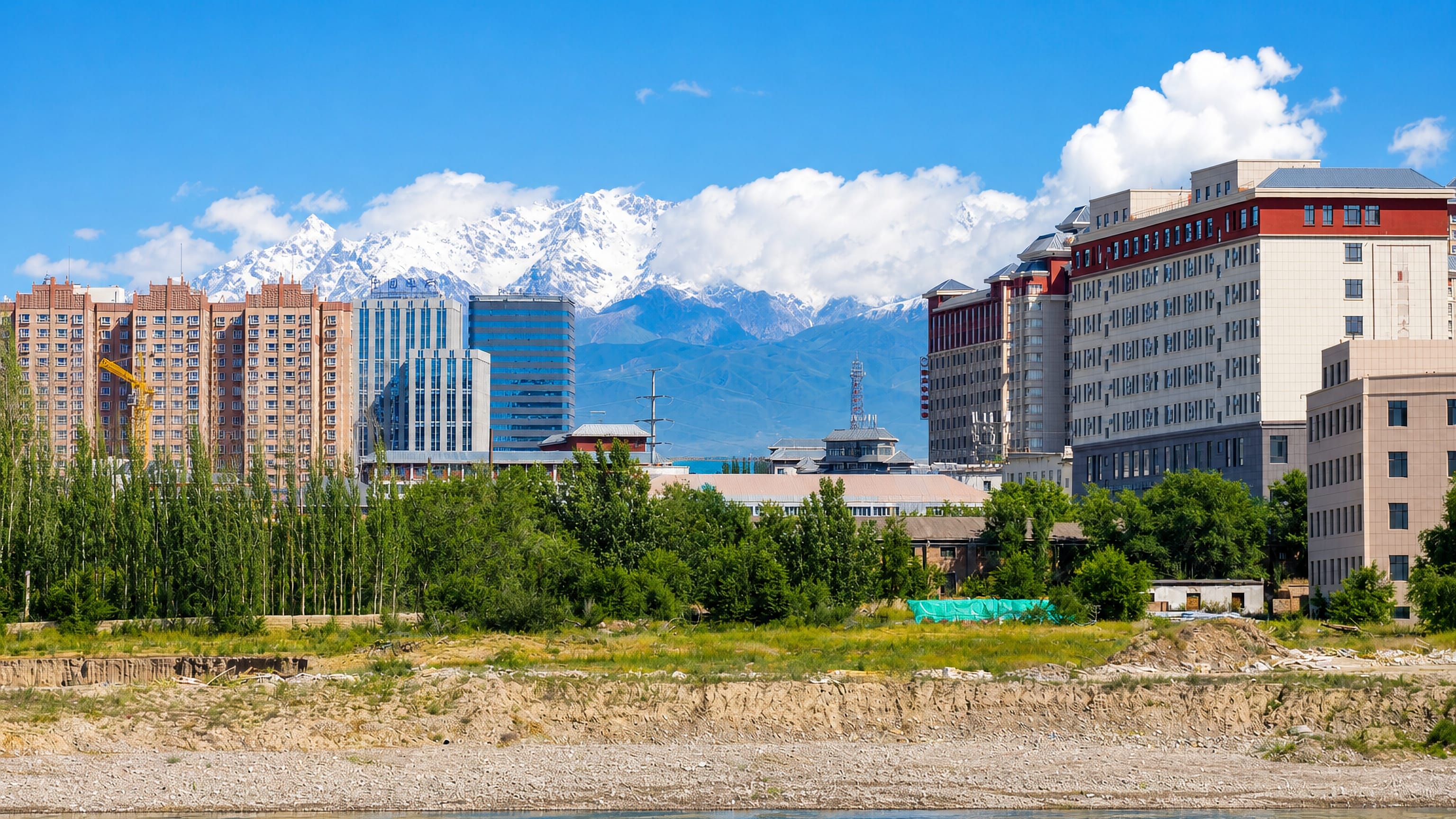 Riverside mid-rise blocks in Qarasu, Xinjiang, backed by the snow-capped Tian Shan under a clear summer sky.
