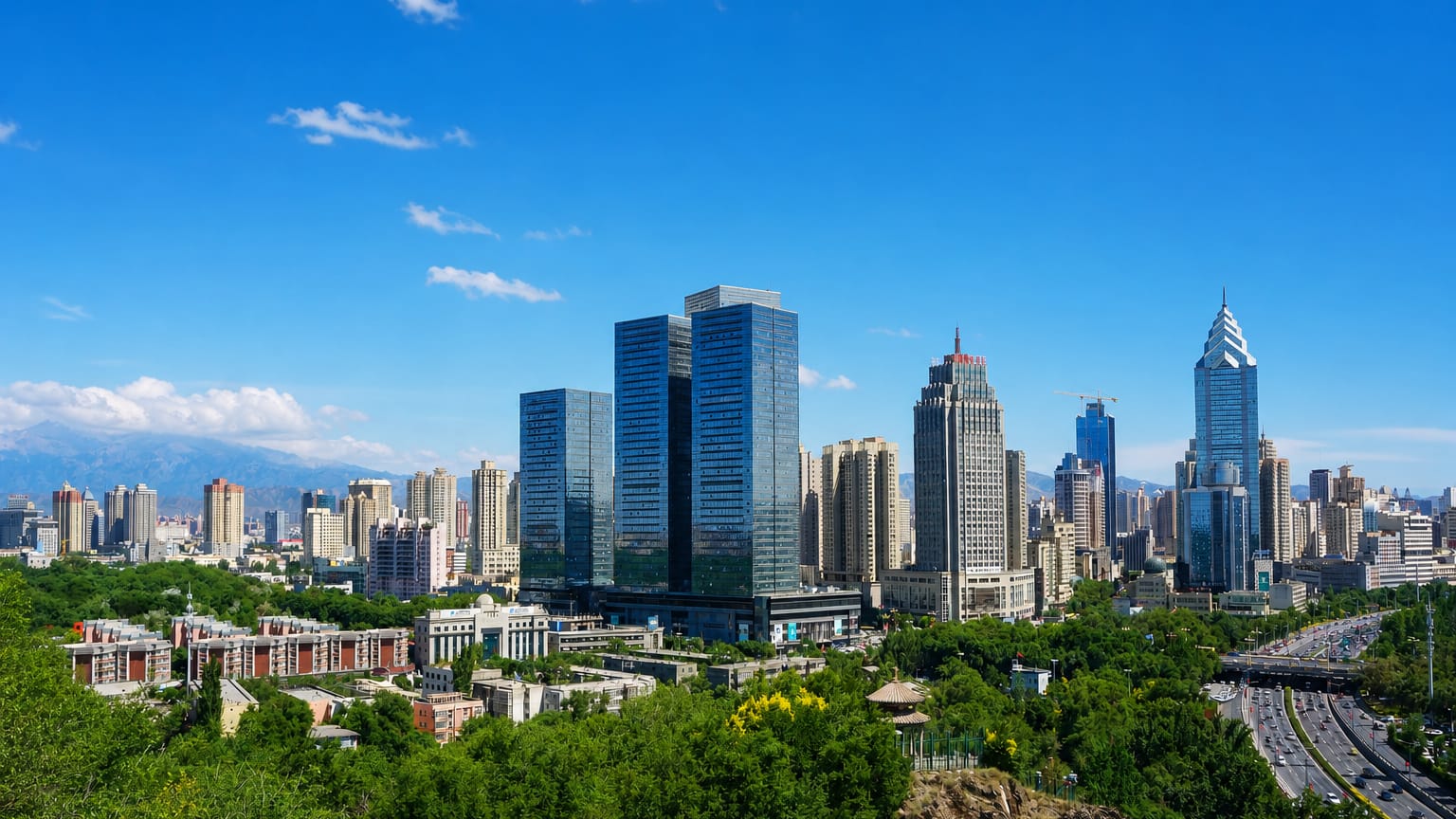 Ürümqi's high-rise downtown with a spire-topped tower and the Tian Shan mountains behind a green ridge.