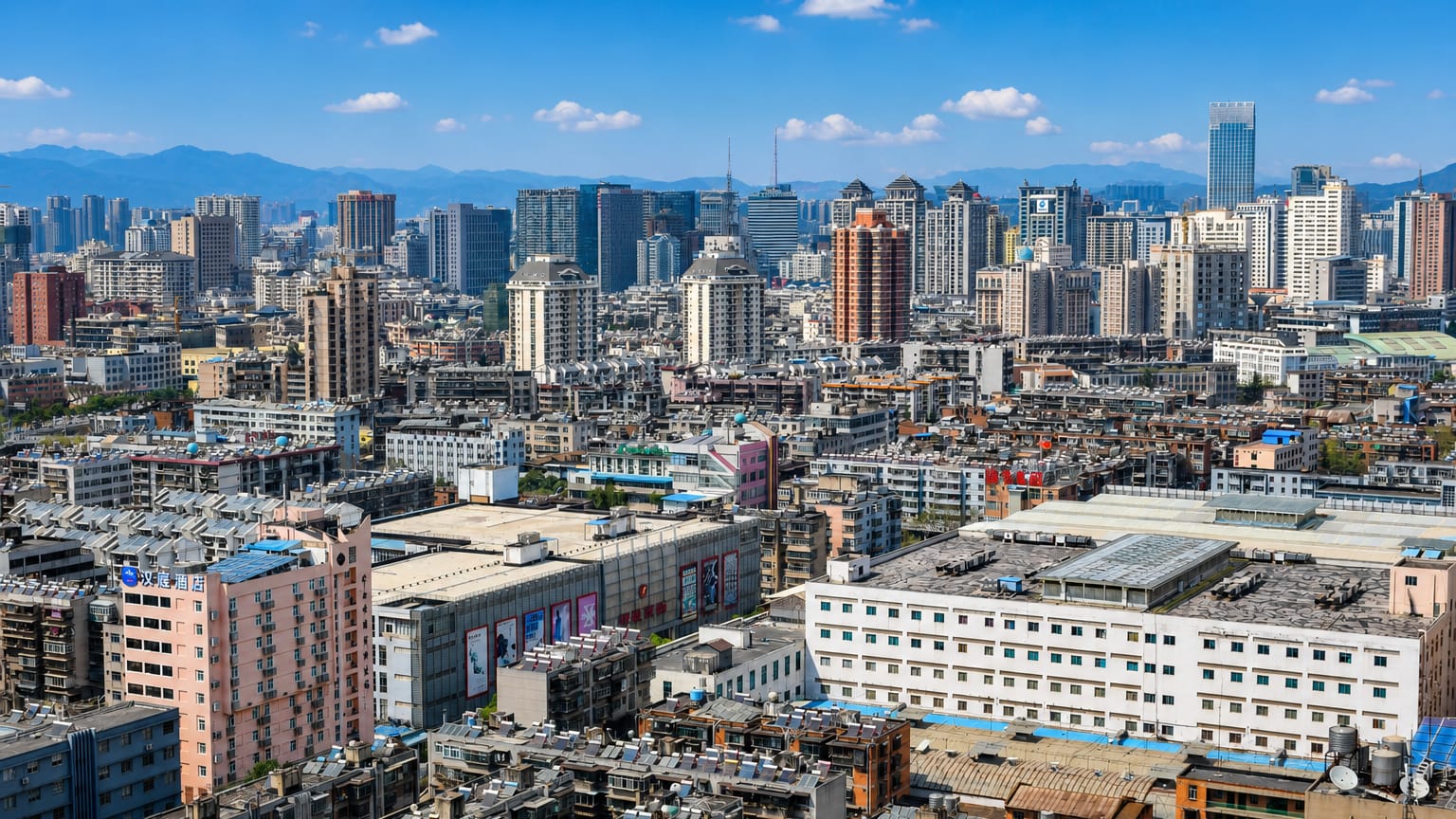 Dense rooftops and high-rises of Kunming stretching toward mountains under a blue sky.