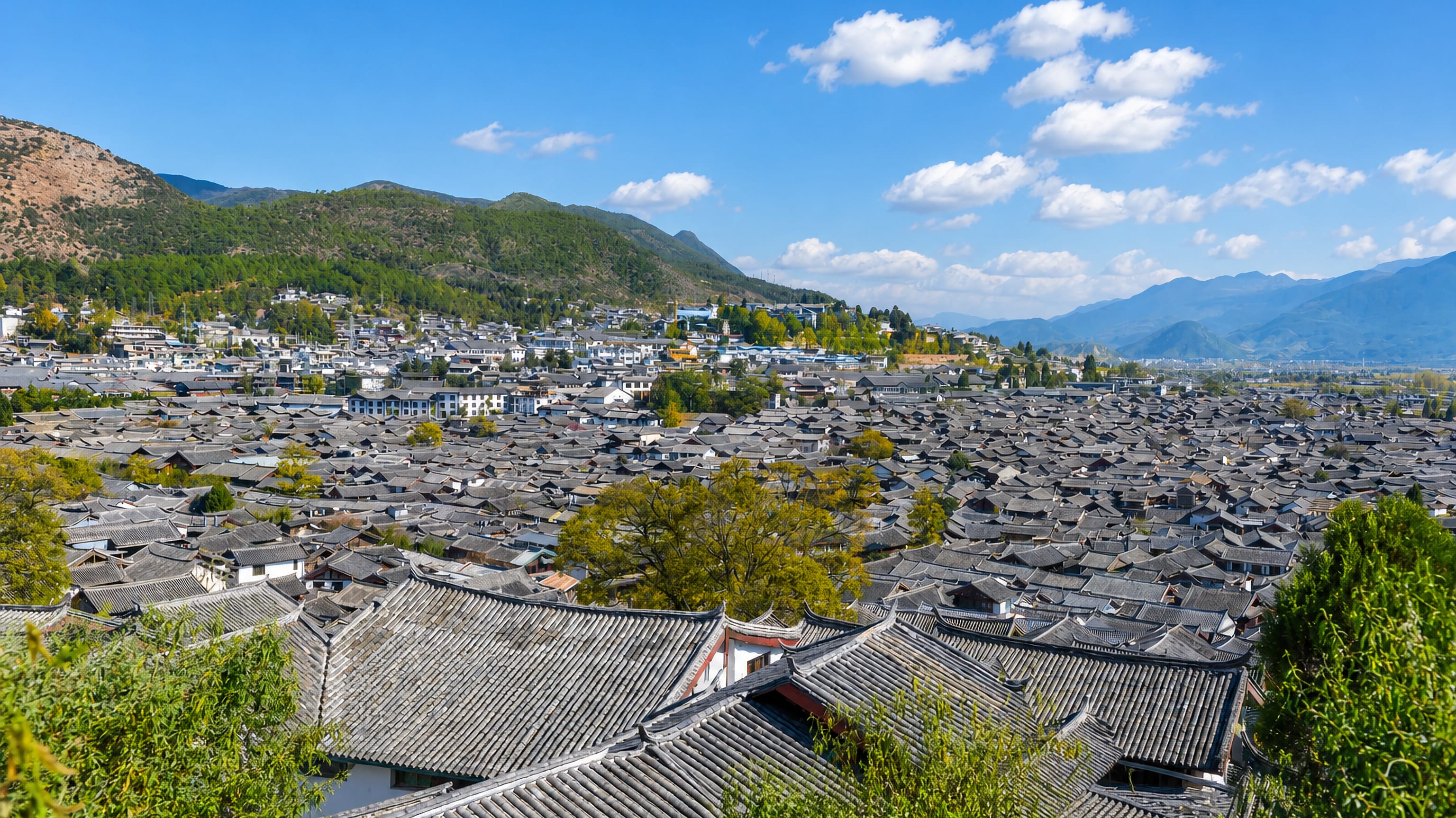 Grey-tiled rooftops of Lijiang Old Town in Yunnan, ringed by wooded mountains under a clear sky.