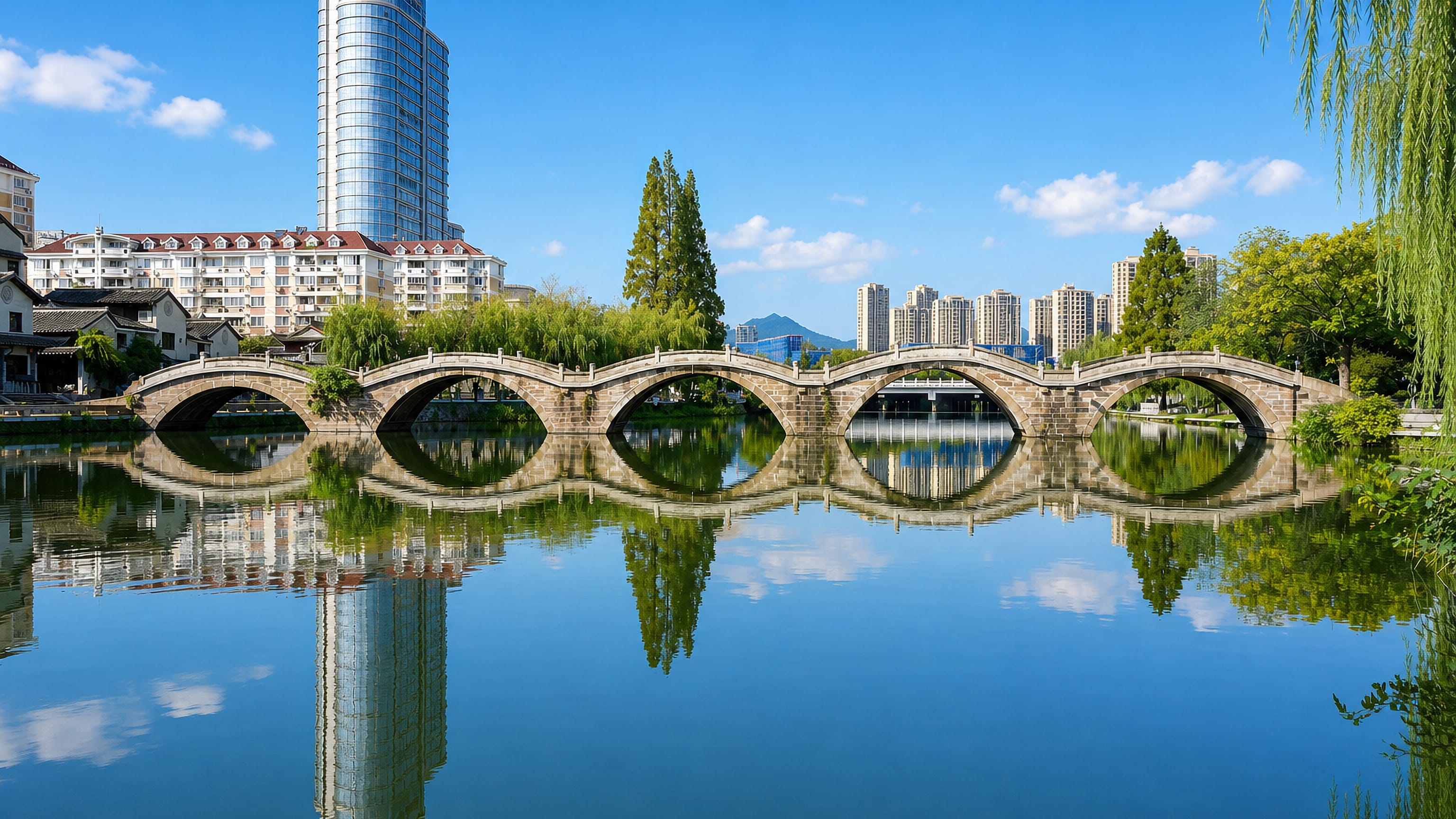 Multi-arch stone bridge mirrored in calm water with modern high-rises behind in Taizhou, Zhejiang.