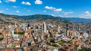 Aerial view of downtown Medellín high-rises ringed by the green Aburrá Valley mountains under a blue sky