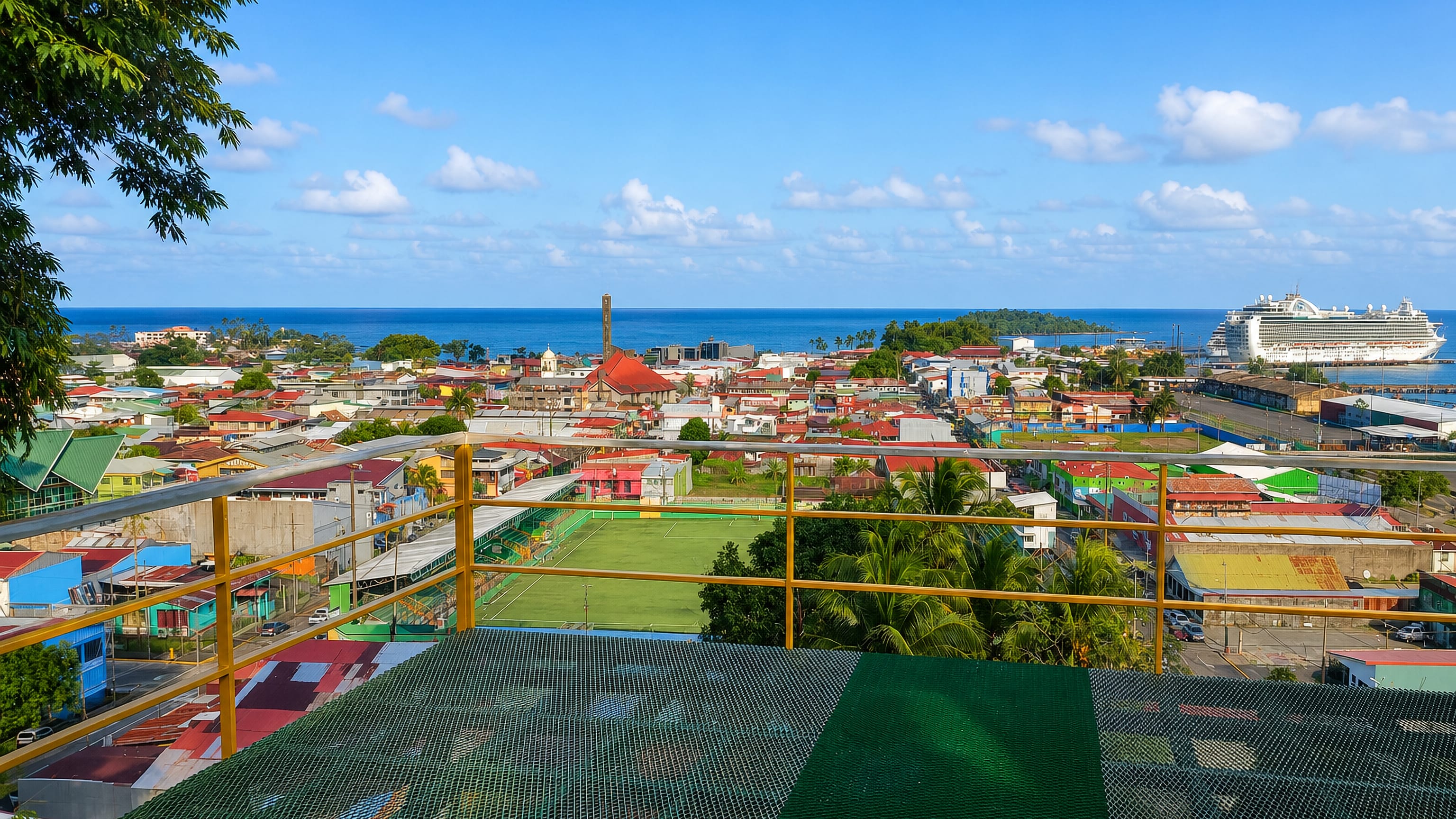 Hillside view over Puerto Limón, Costa Rica, with coloured roofs, a football pitch and a cruise ship at the pier.