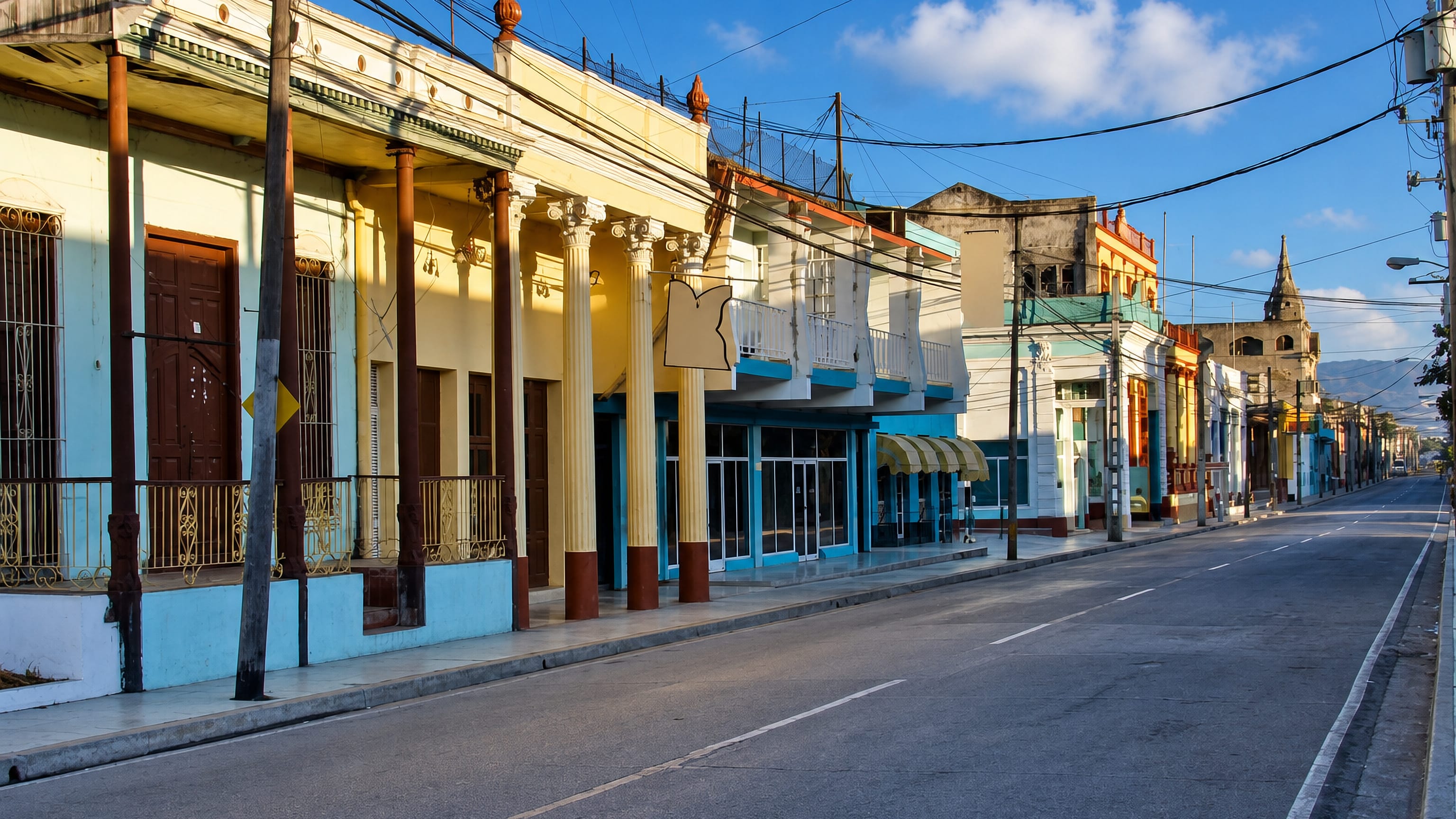 Colonnaded pastel storefronts line a quiet street in Guantánamo, Cuba, with a church tower and hills beyond.