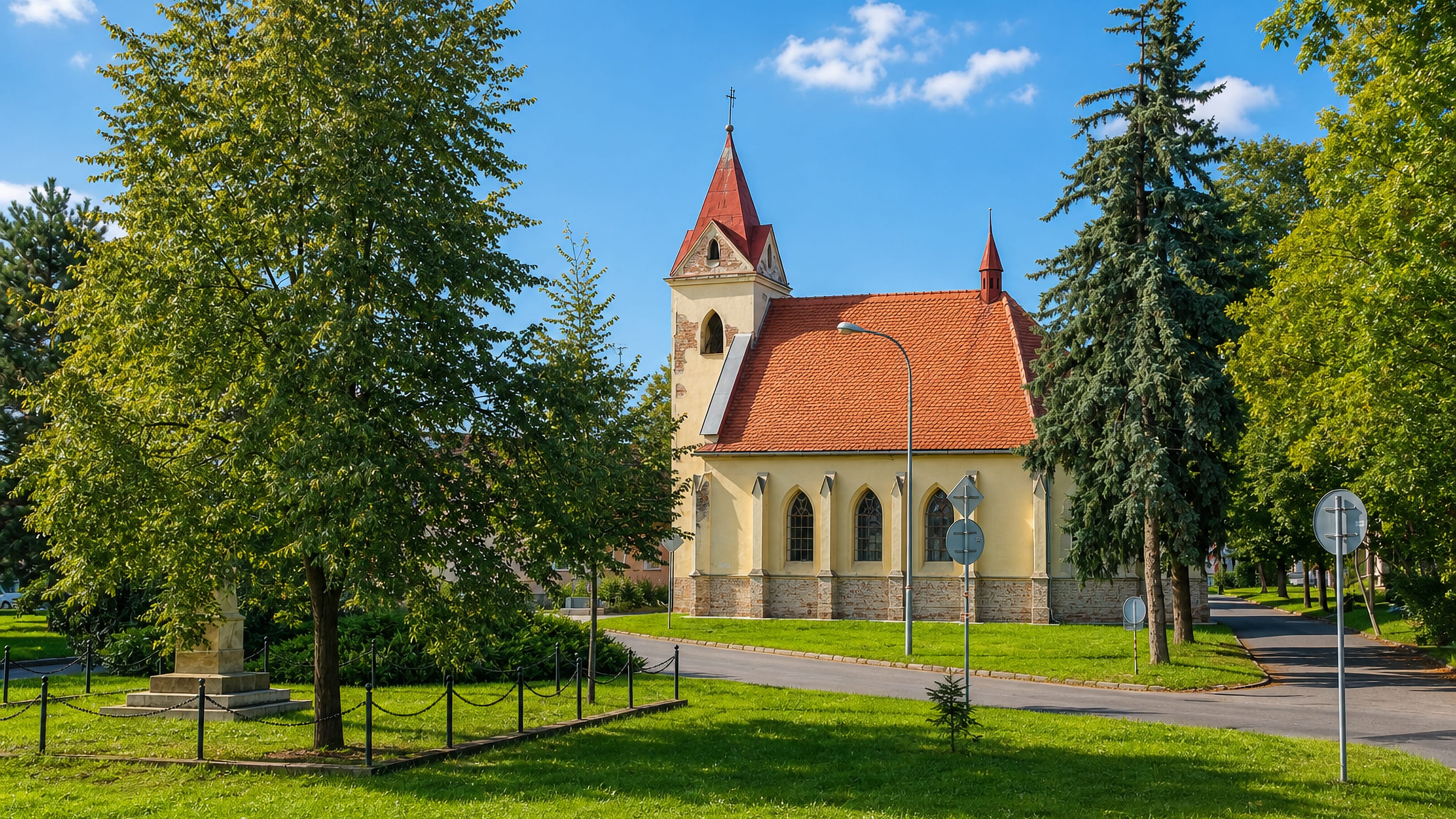 Small yellow village church with a red-tiled roof and a memorial on the green in Prizrenice, Brno.